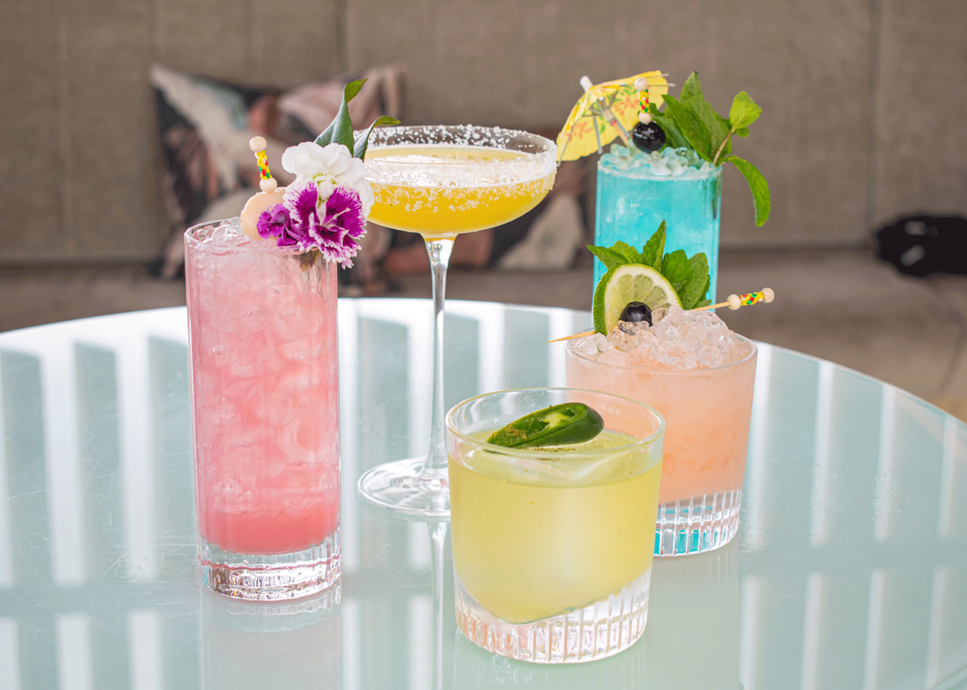 Five bright and colourful cocktails with garnishes on a glass table.