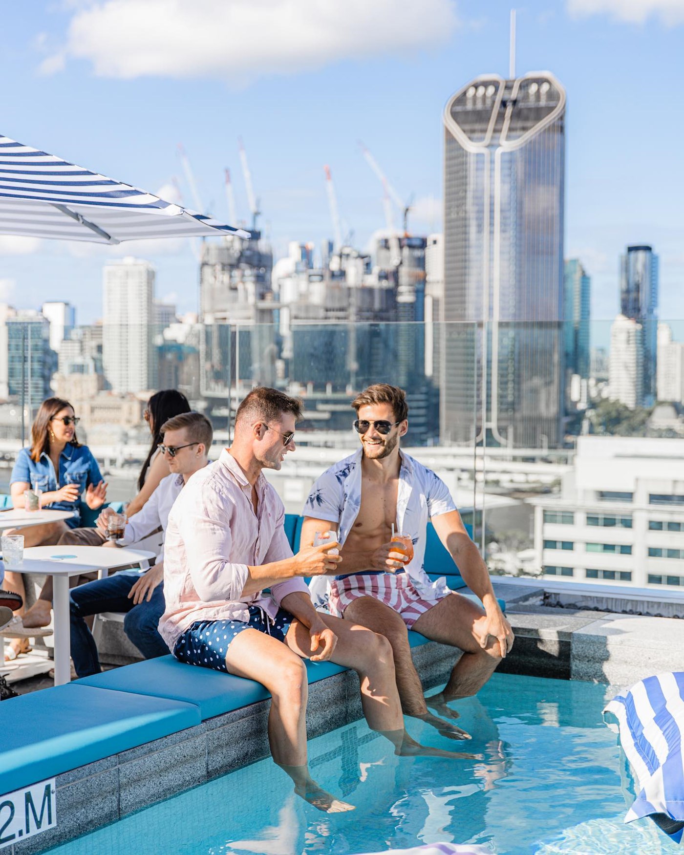 Two people sit on the edge of pool at a busy rooftop bars, with their feet in the water. 