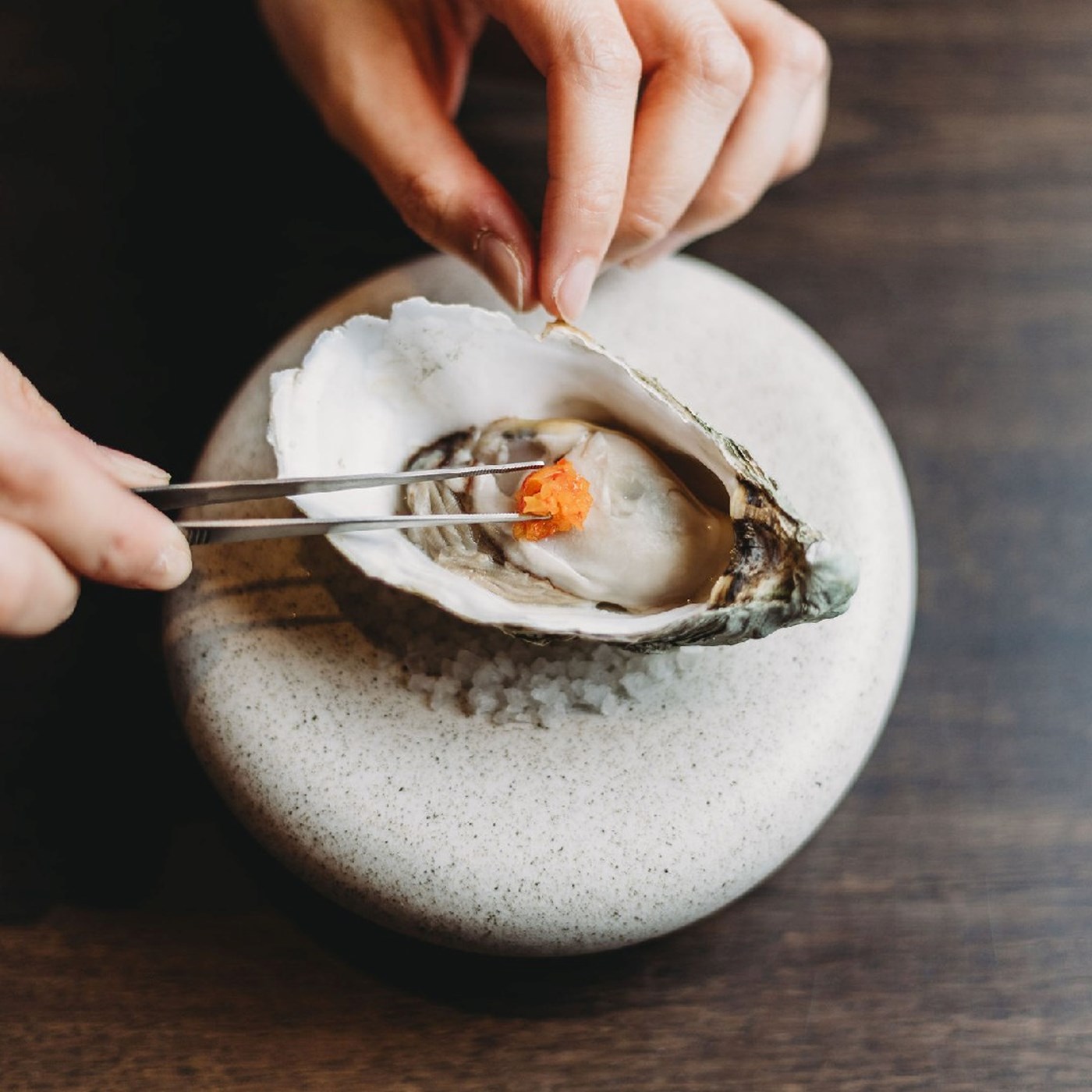 A single oyster sitting atop a stone dish being garnished with a pair of chef tweezers 