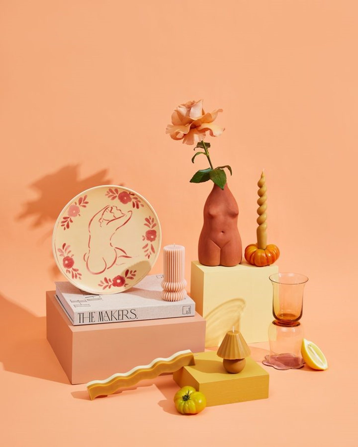 An apricot background with an array of homewares including wiggy candles, a woman silhouette vase and a plate. 