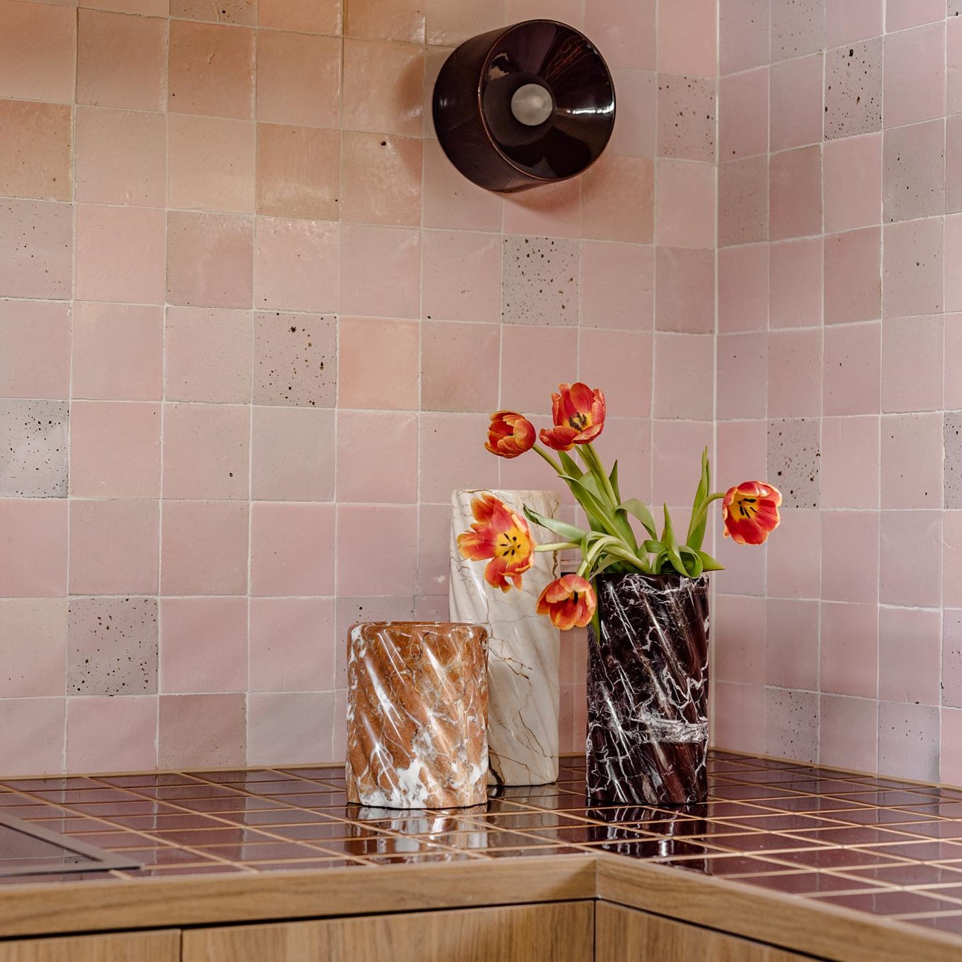 A pink tiled kitchen with three marble vases from Paolo & Joy in the corner, one of which is holding some orange tulips 