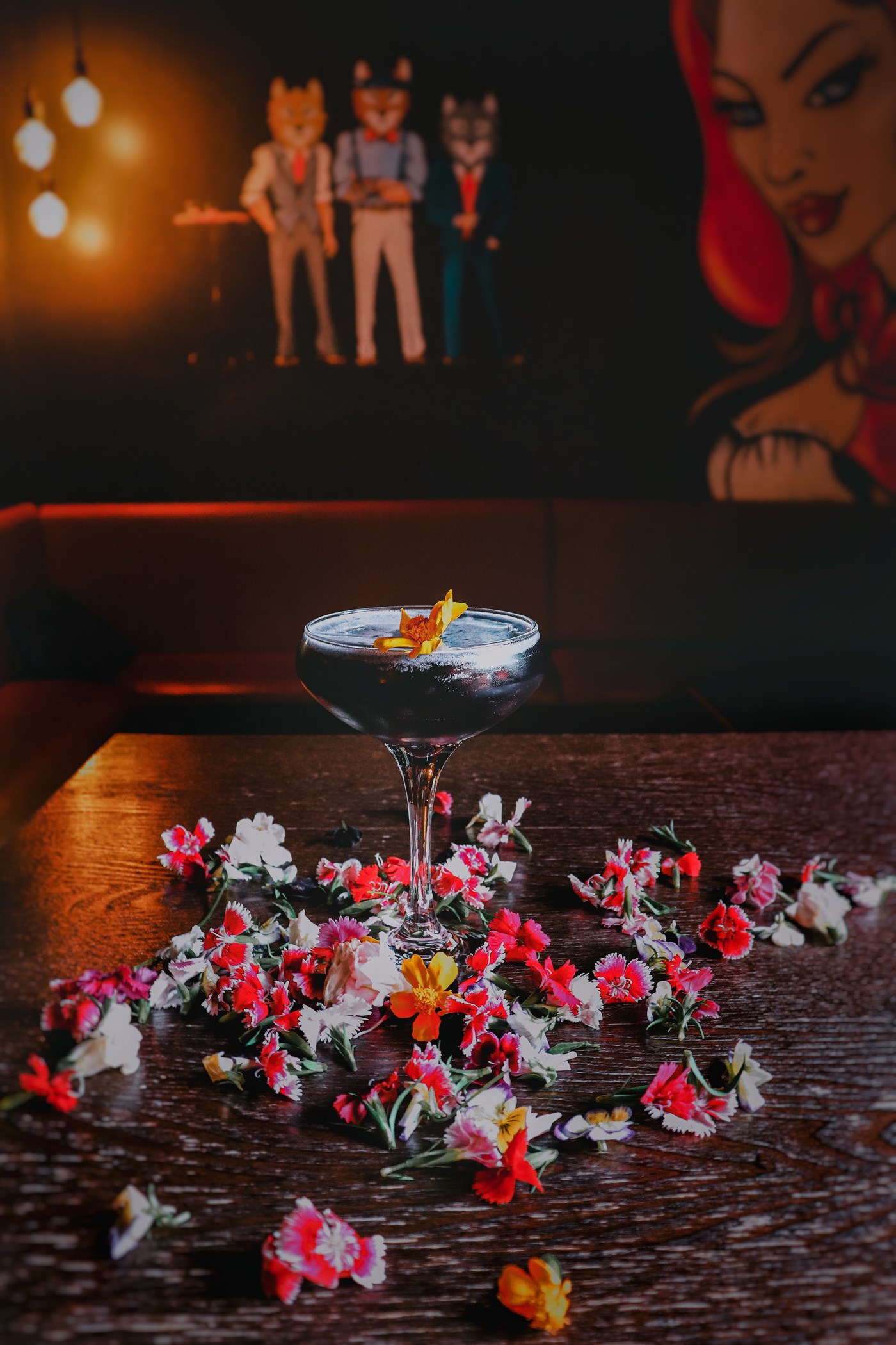 A dark cocktail sits on a brown table, surrounded by scatted garnish flowers.