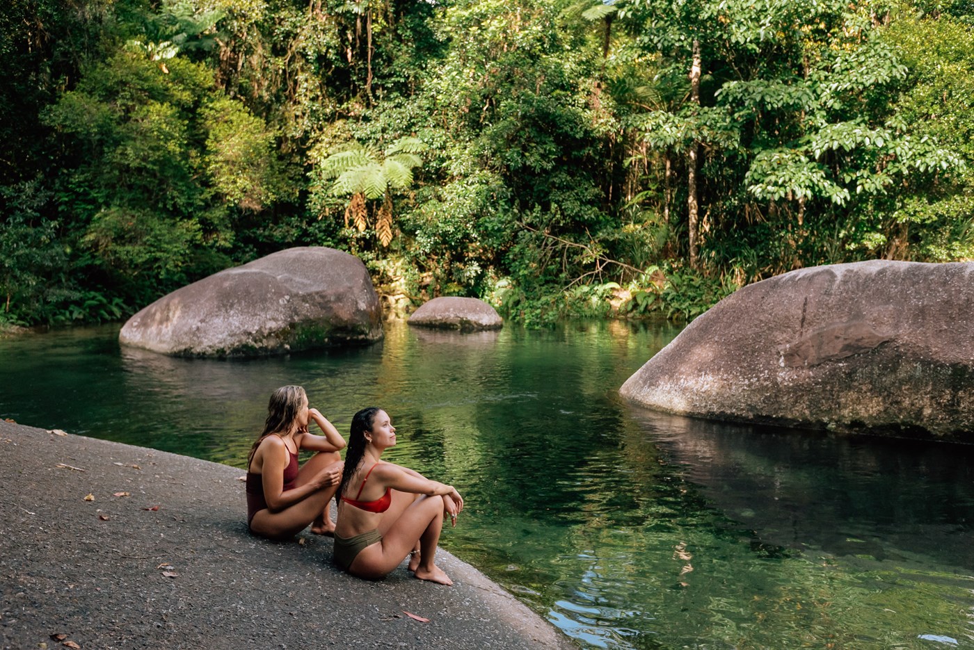 Two people sitting on a large rock gazing into the water, surrounded by leafy dense rainforest