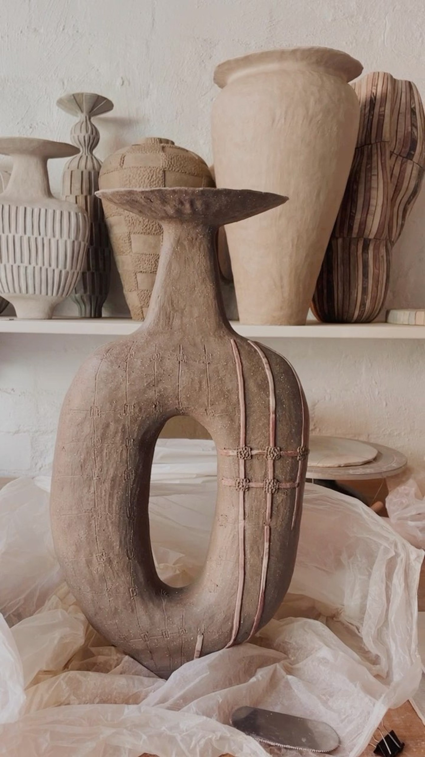 A large raw clay vessel with a hole in the centre and a fanned out top. Other similar vessels are visible in the background 