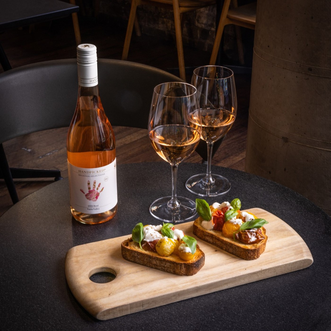 Two glasses of wine sitting next to a bottle and behind a wooden tray of bruschetta. 