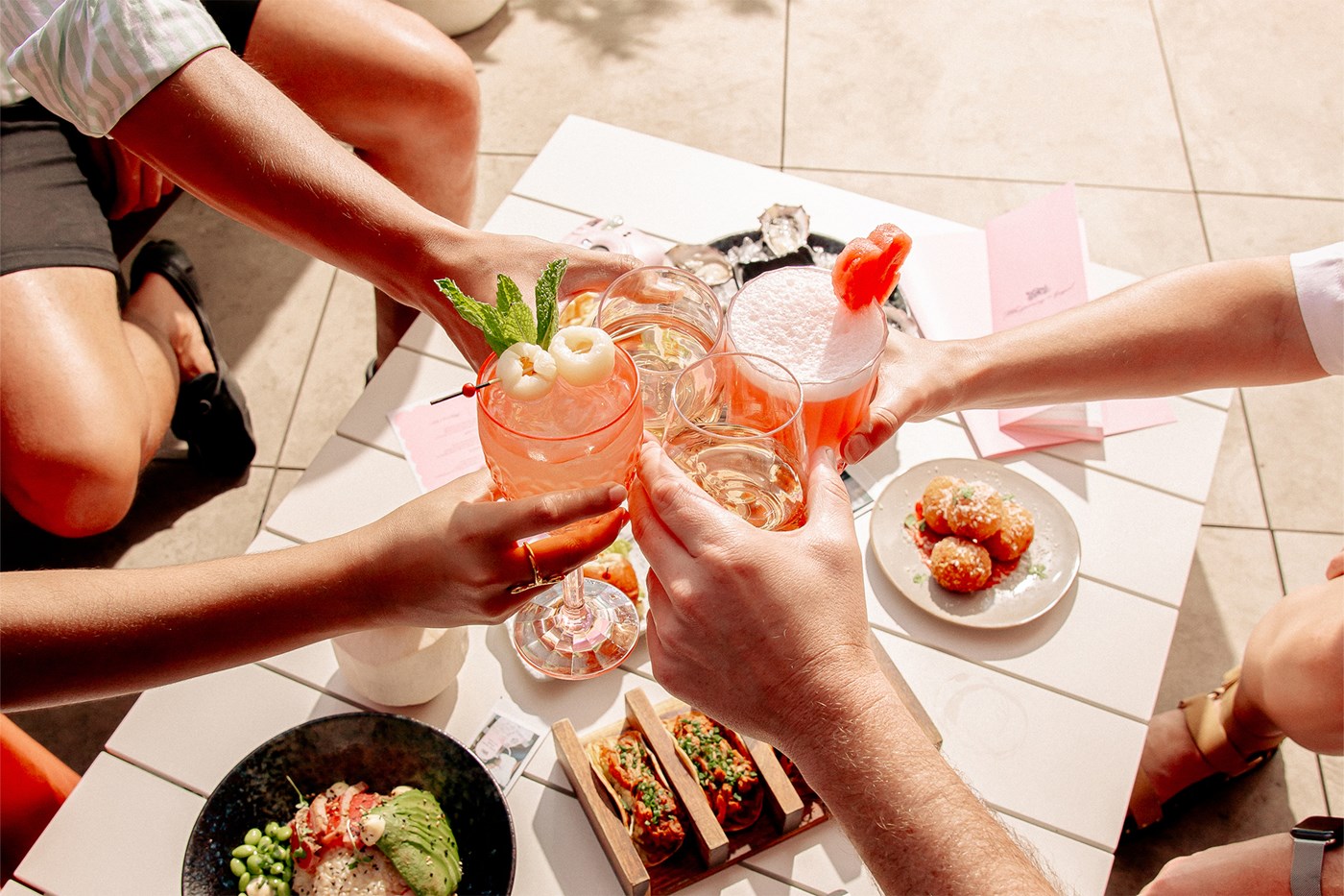 Four people clink pink cocktails together over a white table spread with food.