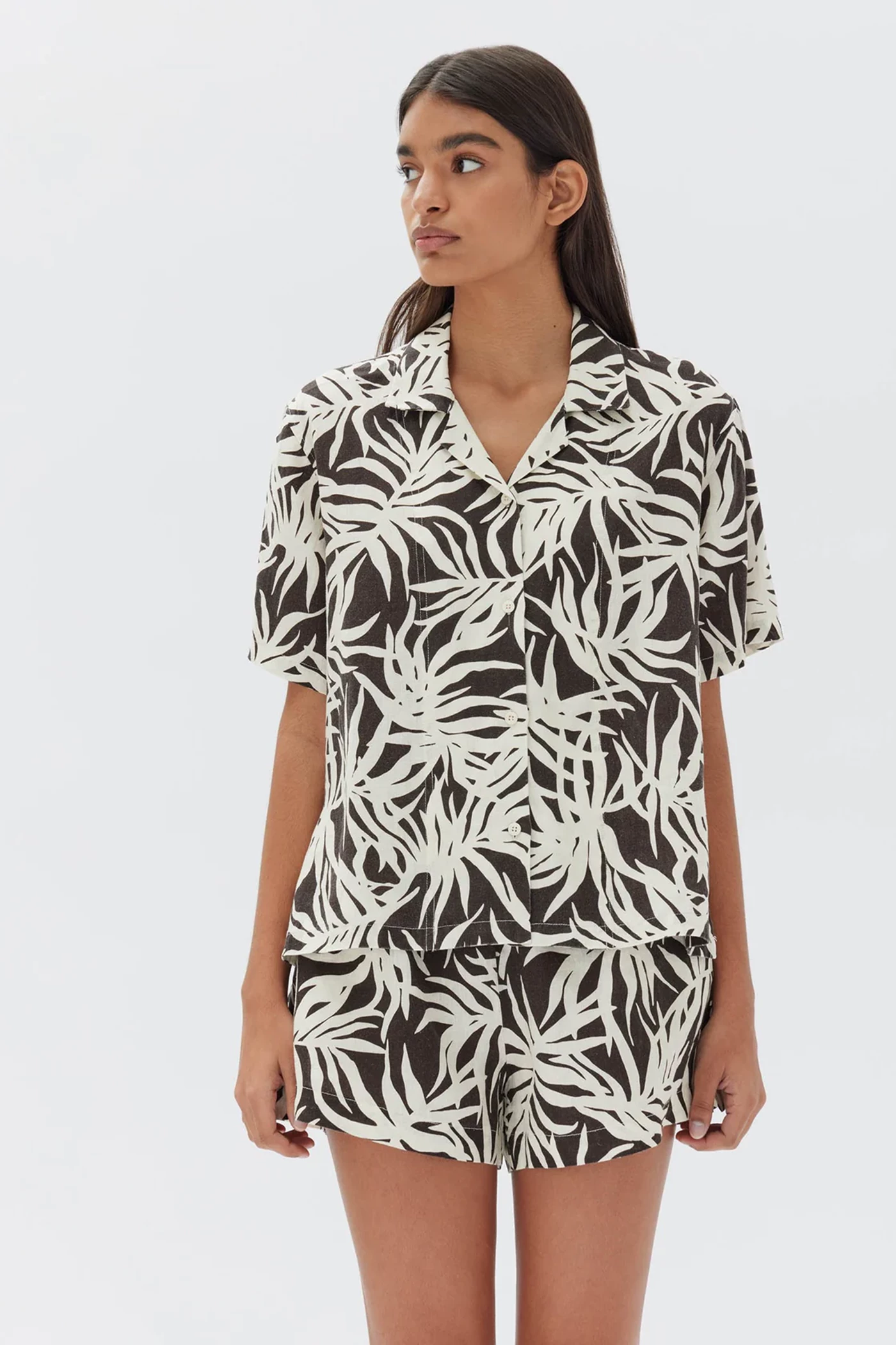 A woman wears a matching linen set in a short-sleeve shirt and shorts, both in a palm tree print.