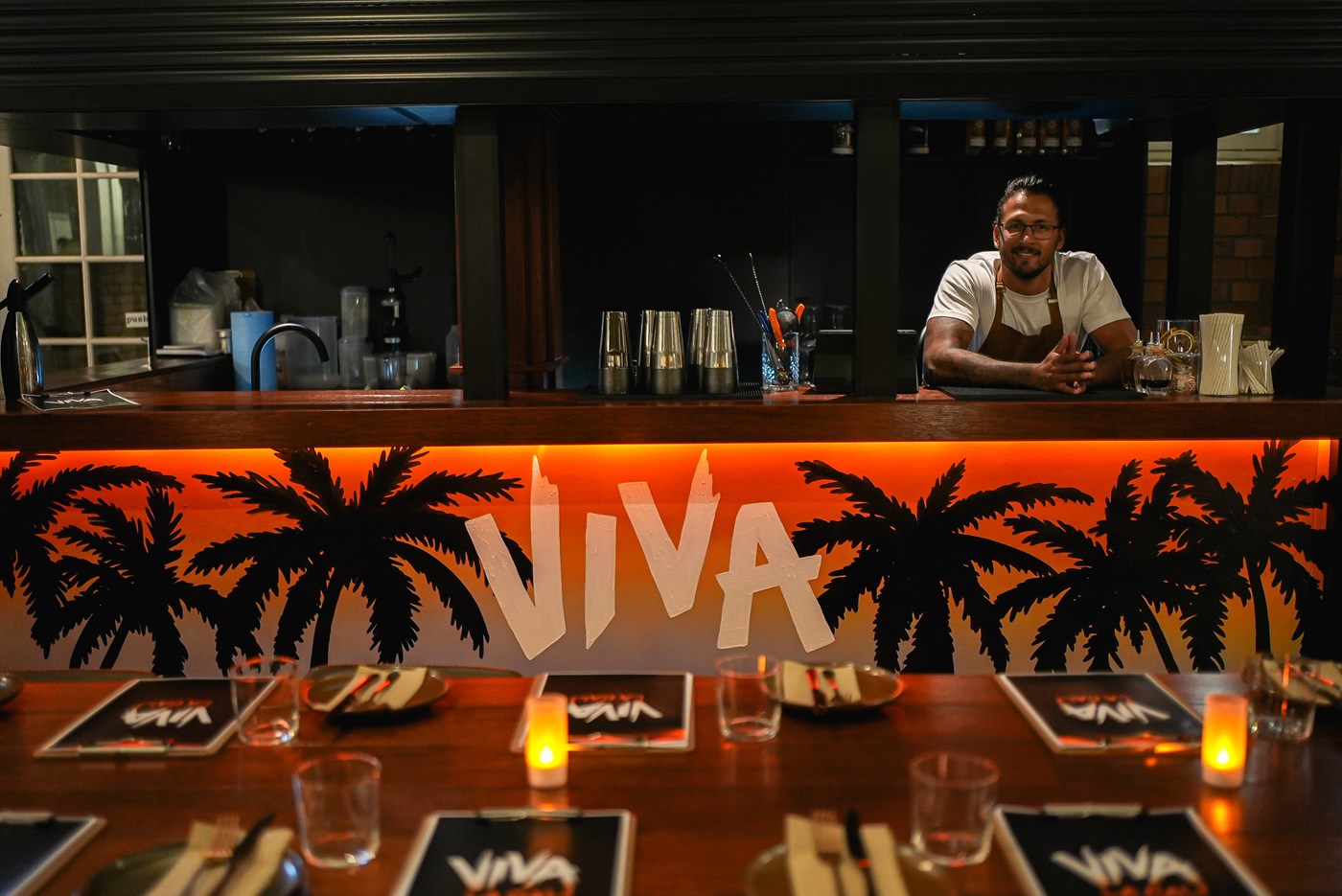 A dimly lit bar with "VIVA" and palm trees painted on the front. The bartender is smiling & leaning on the bar 