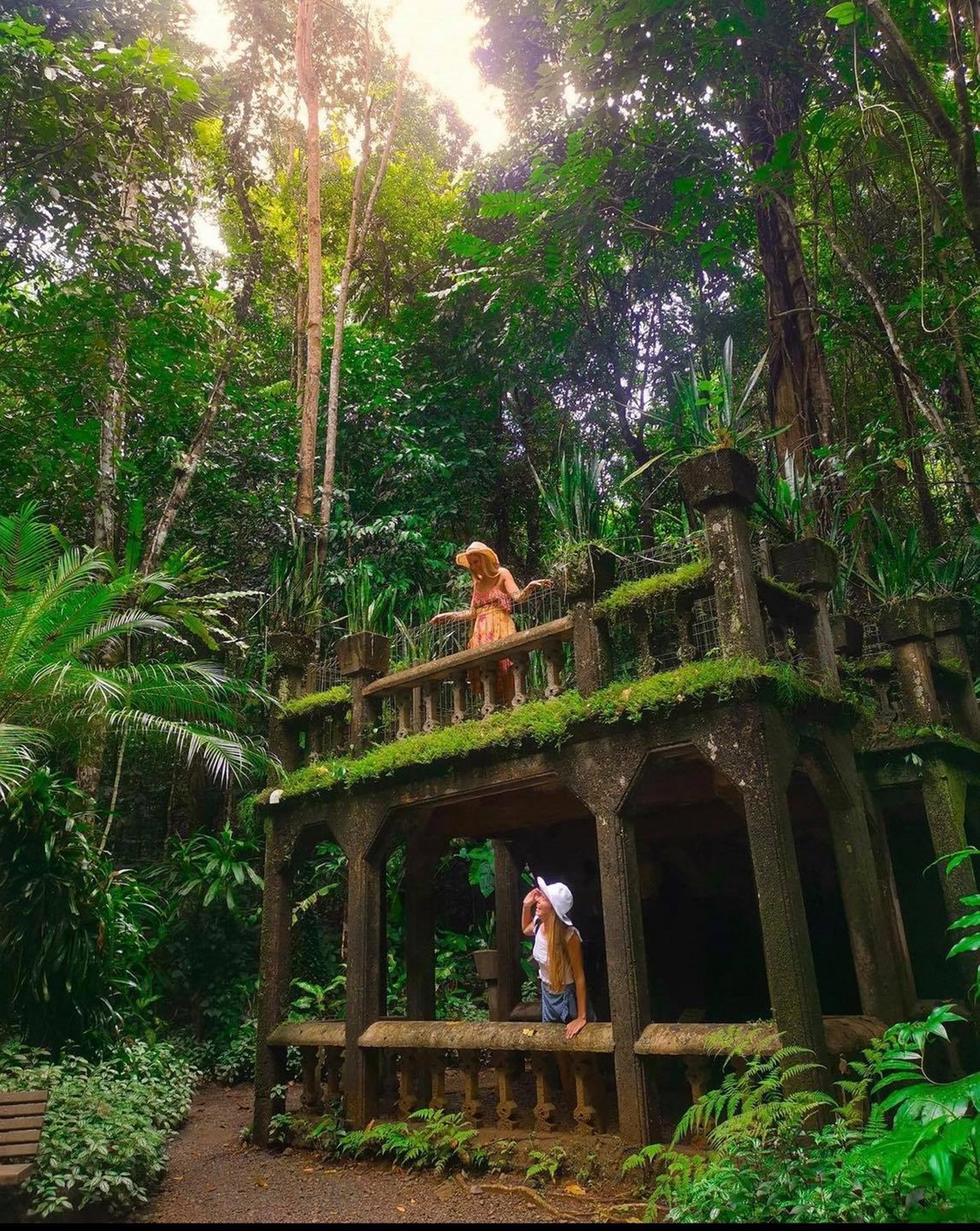 An old ruined stone building in a rainforest, one person is on the top balcony level and another person is looking up at them