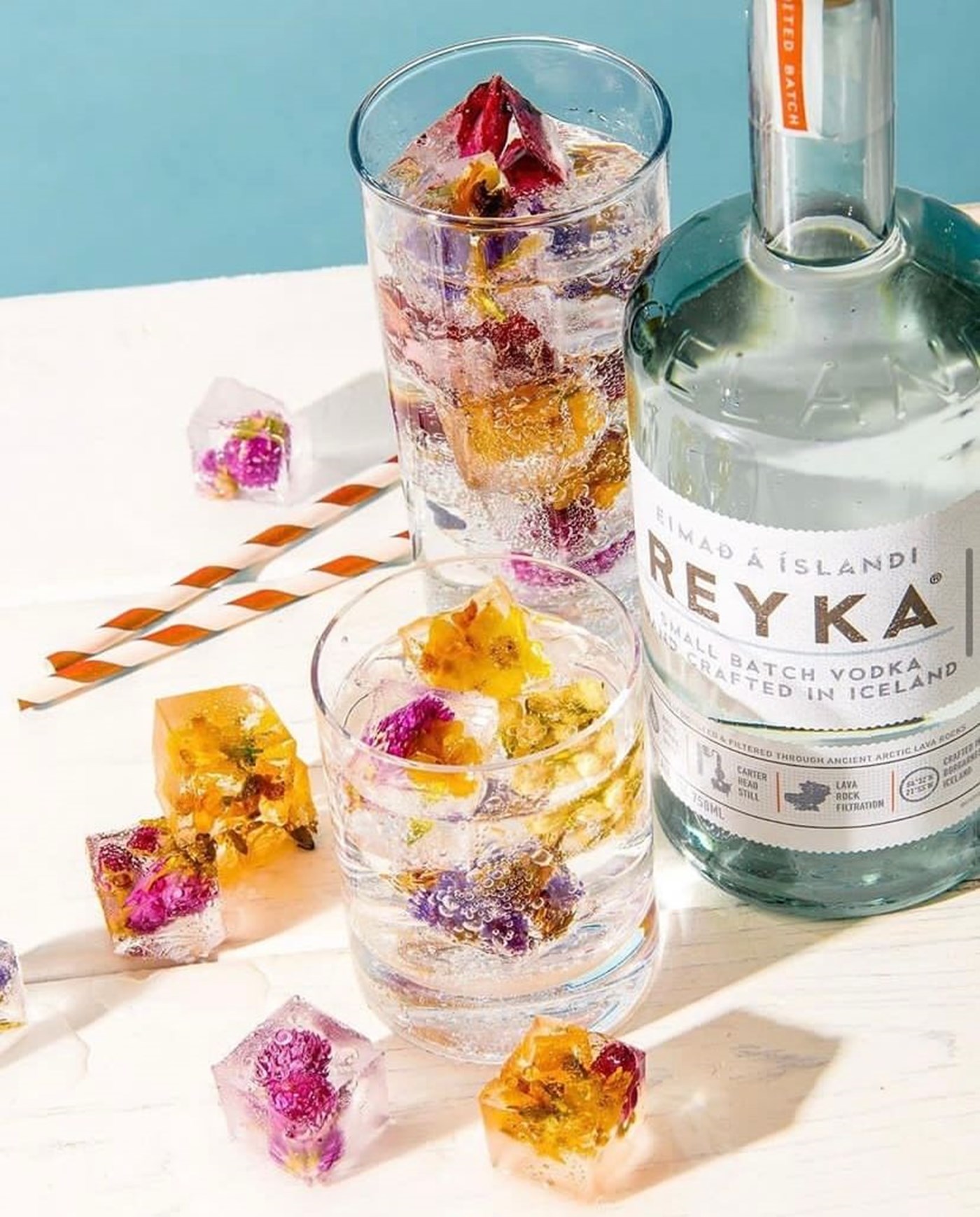 A bottle of reyka vodka next to a mixed drink with flower filled ice cubes spread across the table