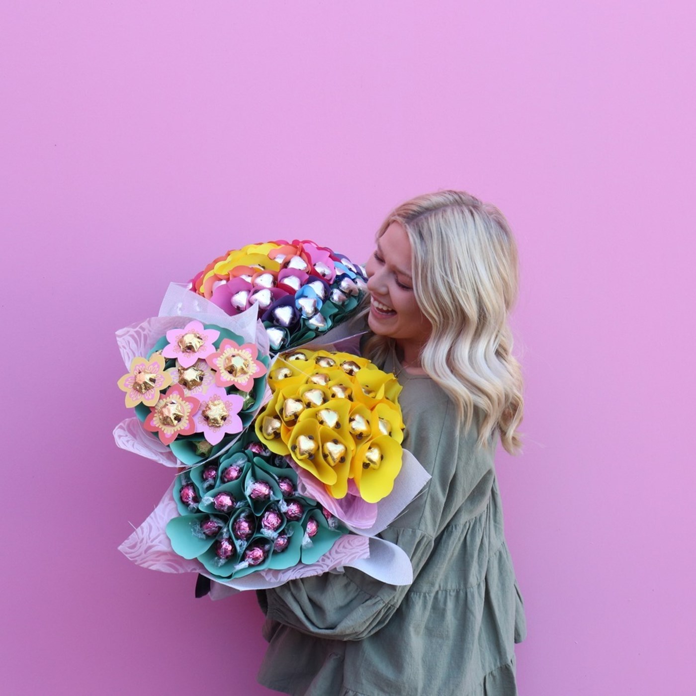 a blonde woman holding up four edible flower bouquets against a plain magenta wall