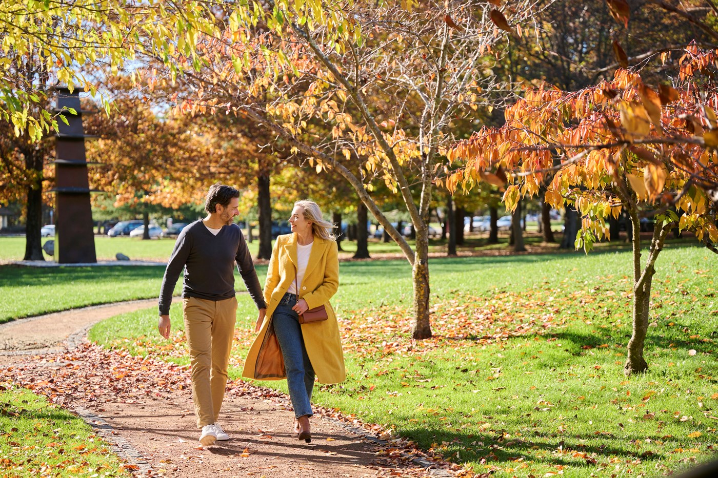 Woman in yellow coat walks with a man in warm clothes thorugh park with orange and yellow leaves at Nara Peace Park Canberra