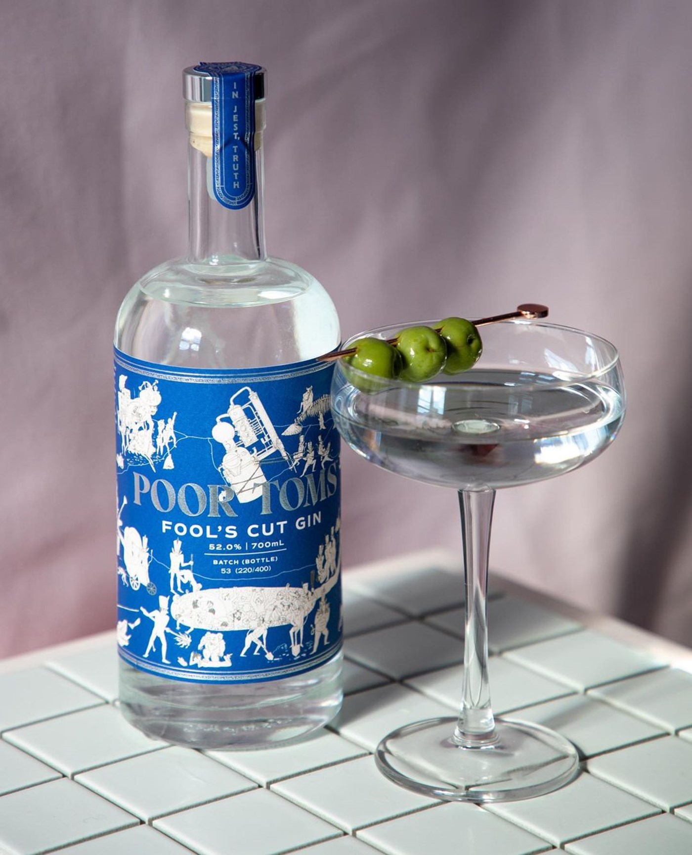 A bottle of Fool's Cut Gin with a blue and white label next to a cocktail in a martini glass atop a tiled bench 