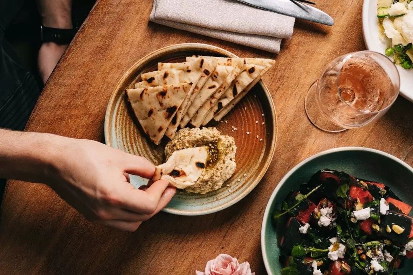A hand dipping a pitta into dip with a salad and glass of rose to the side 