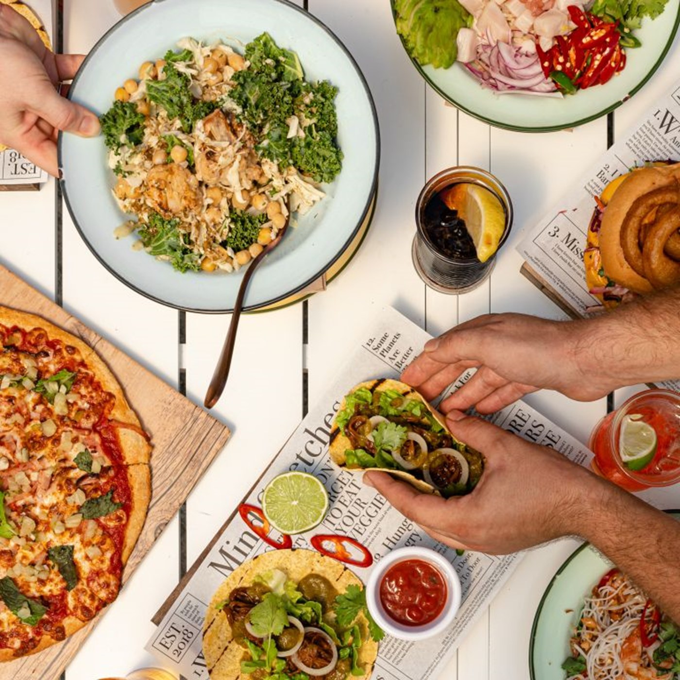 Top down view of a white table with hands reaching for share tacos, pizza and salads
