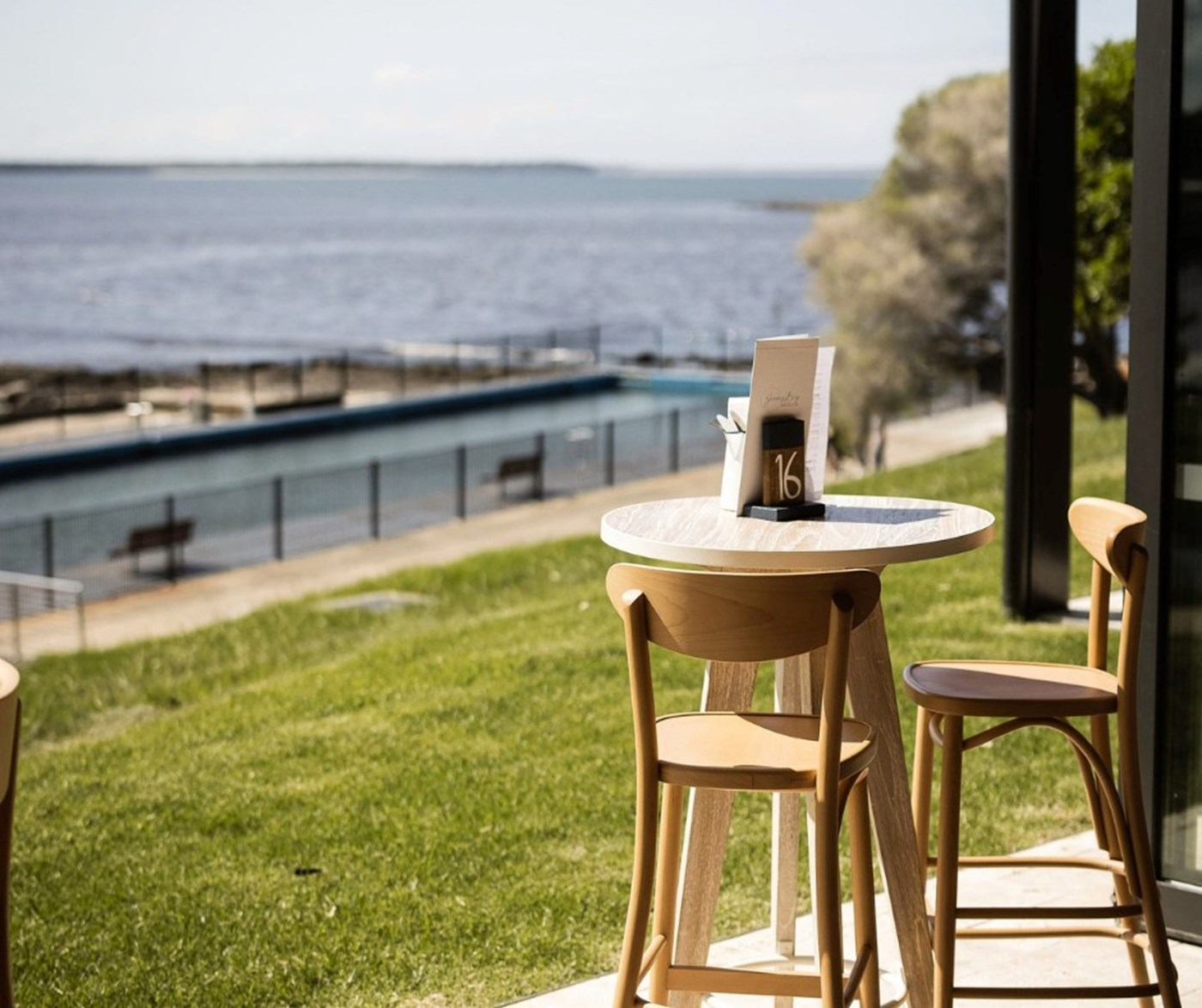 A single table with two chairs overlooking a swathe of grass and an ocean pool 