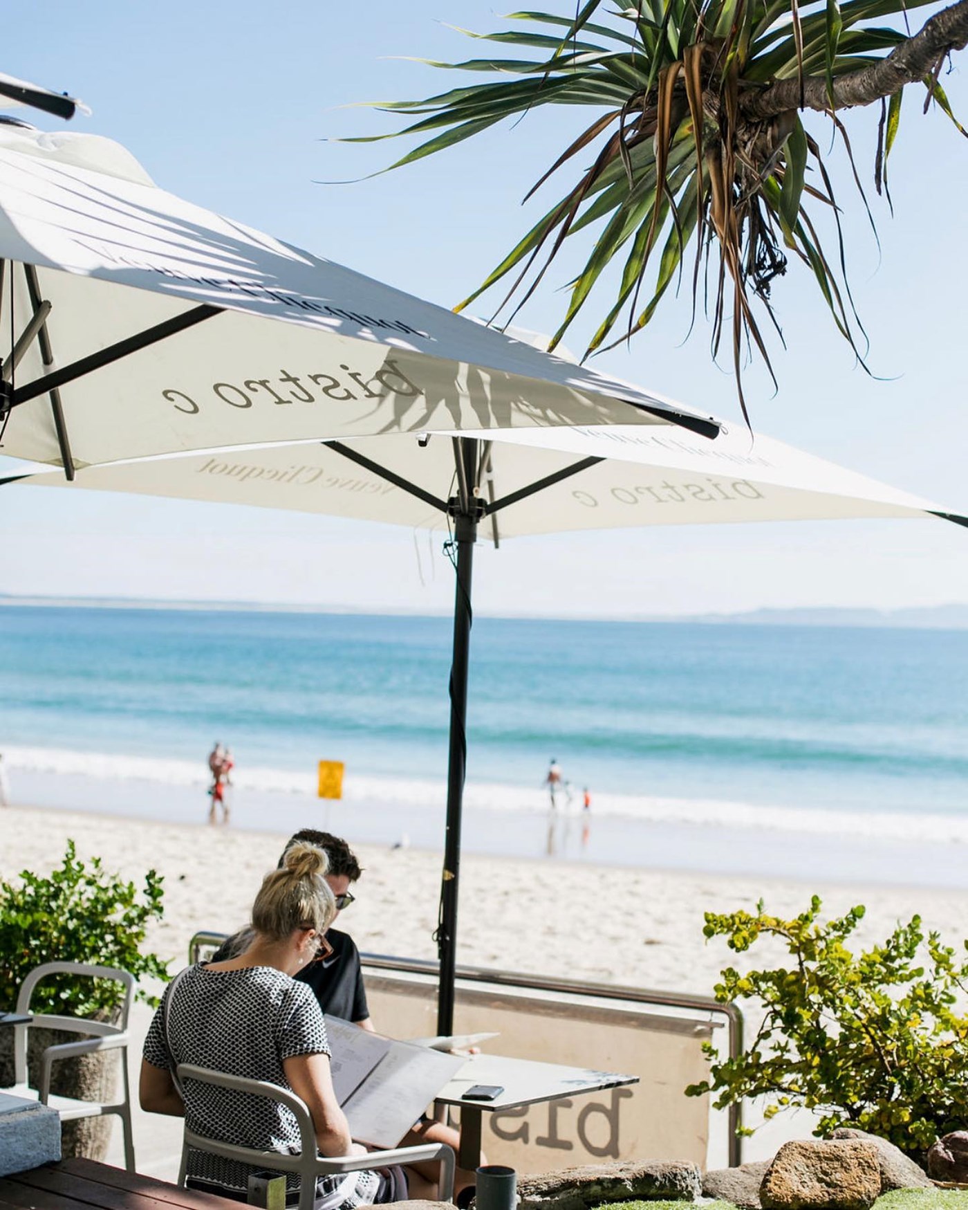 Two people seated at a table under an umbrella are reading the menu, the beach is in the background