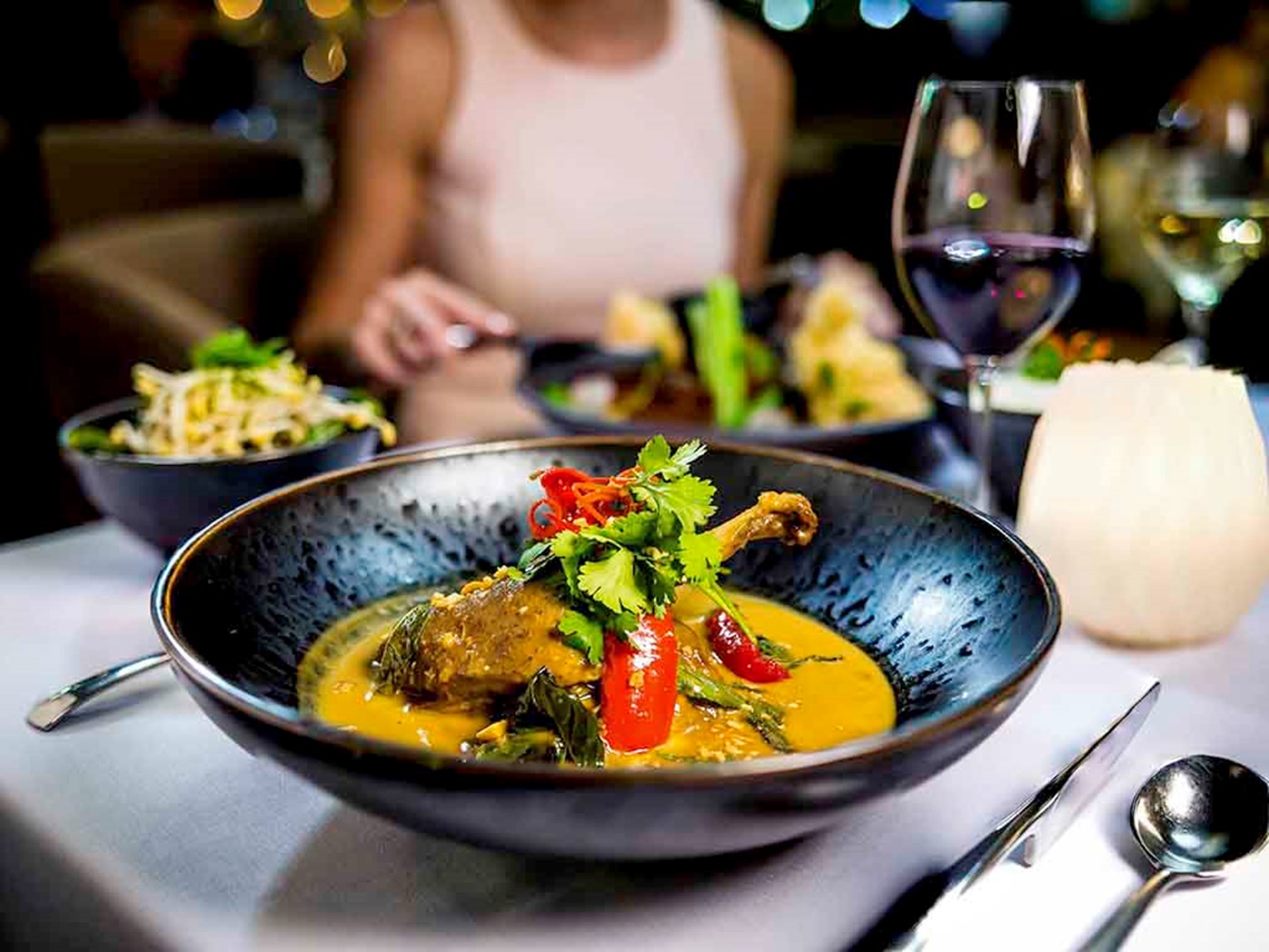 A curry style dish with a quail leg served in a black bowl with a glass of red wine.
