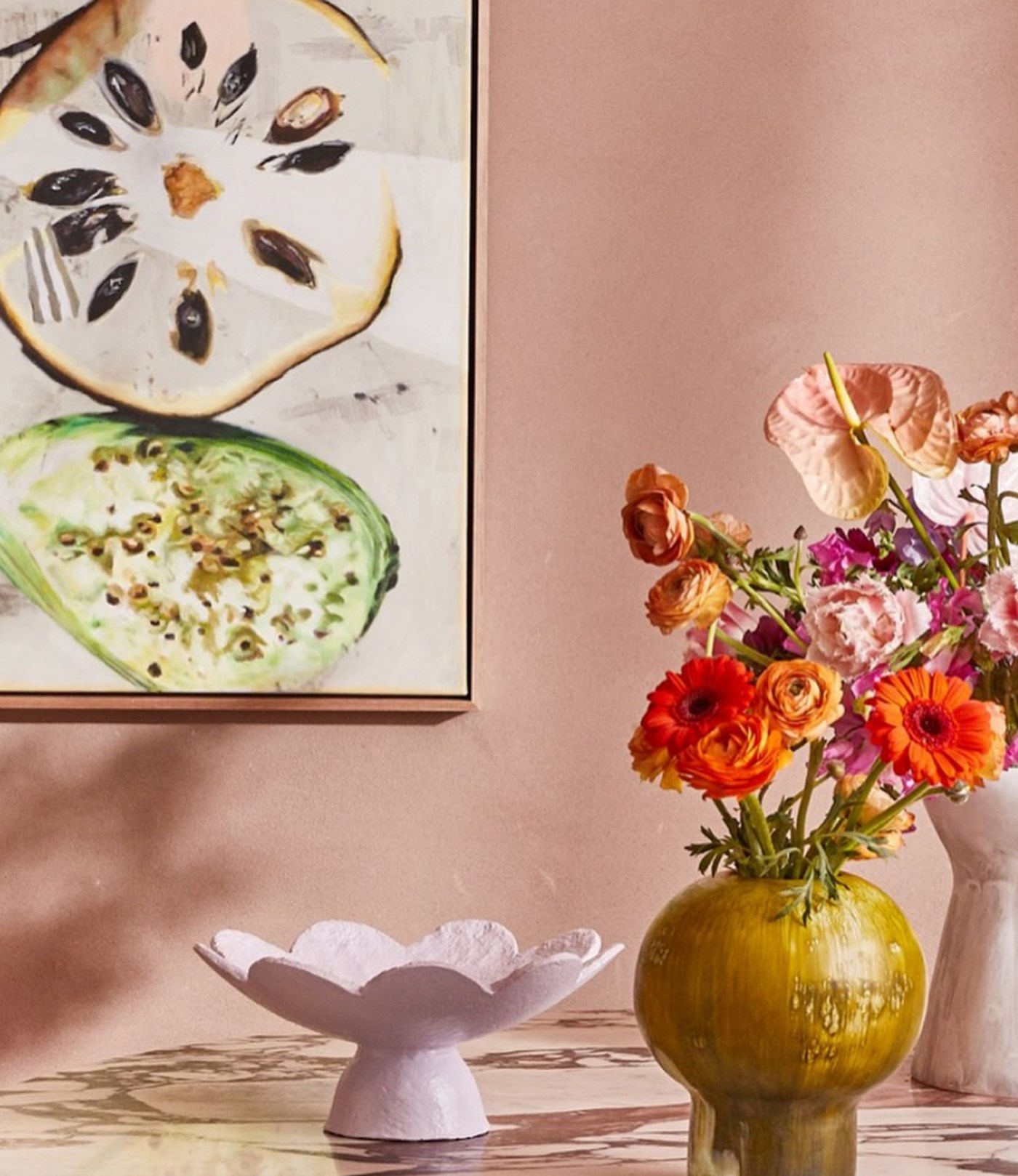 Multiple ceramic objects including a mustard vase full of flowers and a scalloped cake platter against a pink backdrop 