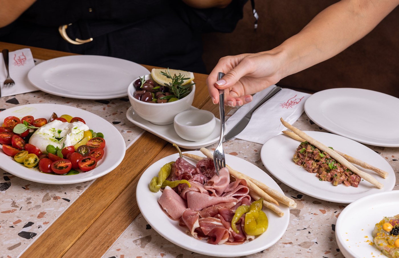 A hand using a fork to dig into a plate of antipasti surrounded by tomato and burrata, breadsticks and olives 