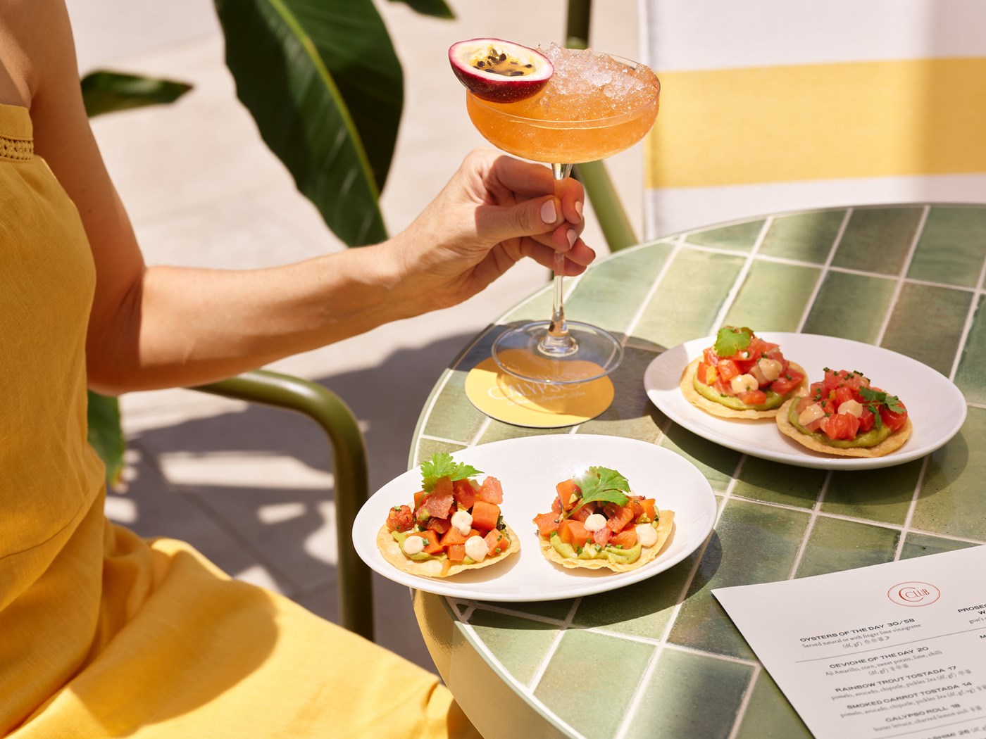 A person in a yellow dress holds a passionfruit cocktail, seated at a green tiled table with two plates of food.