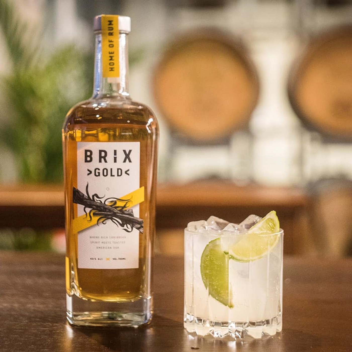 A bottle of Gold Rum from Brix Distillers next to a cocktail on a wooden bench, set against a blurred background 