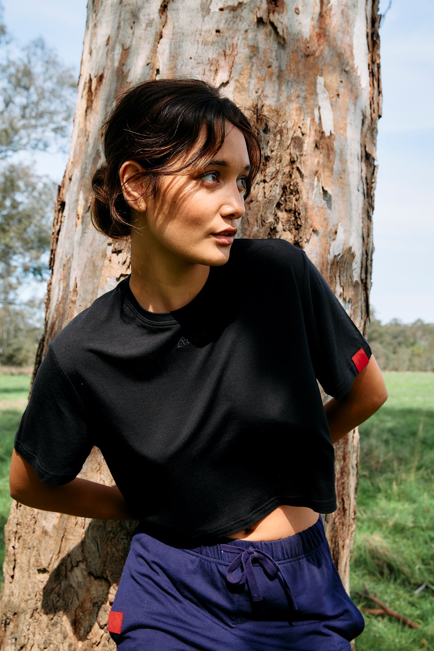A woman wearing a black, cropped t-shirt and blue tracksuit pants leans up against a tree.