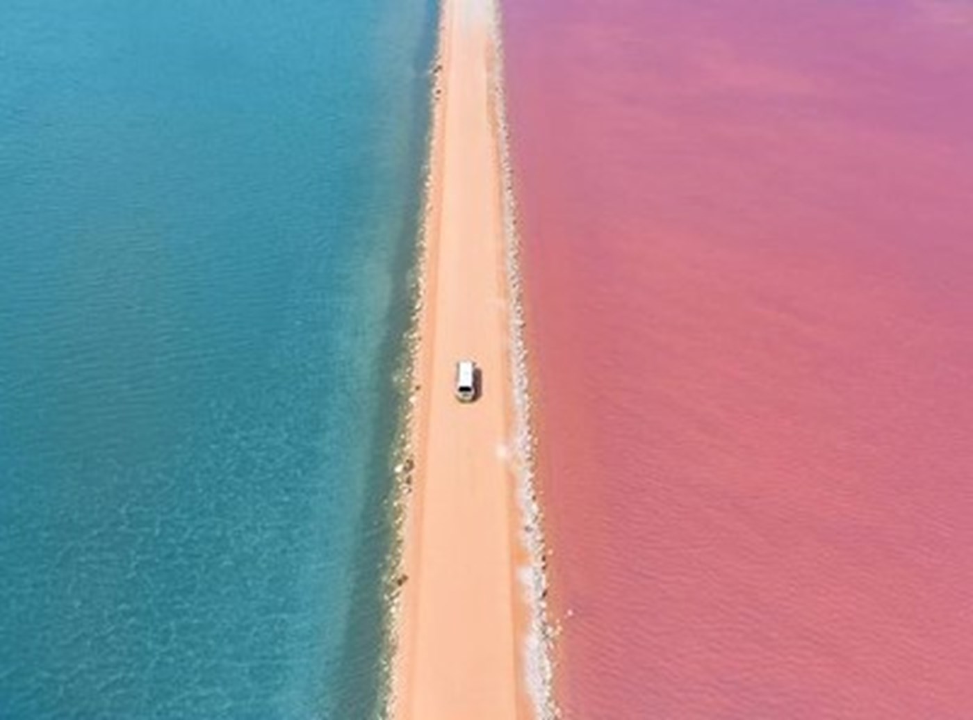 A car drives through a dirt road, framed by a blue lake on one side and a pink lake on the other.