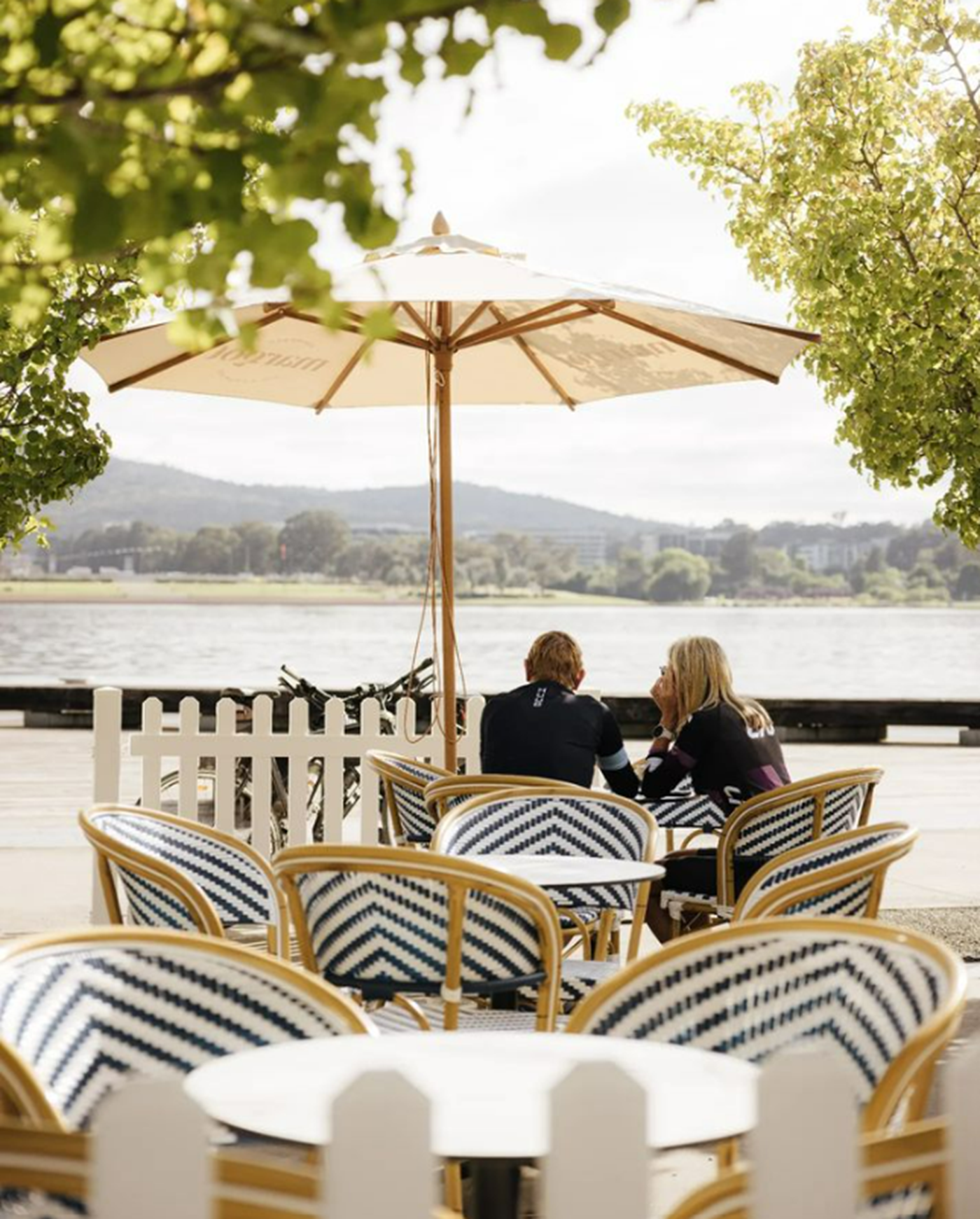 Two bike riders stopping by a lakeside cafe with blue and white cafe seating, taking in the views of the lake 