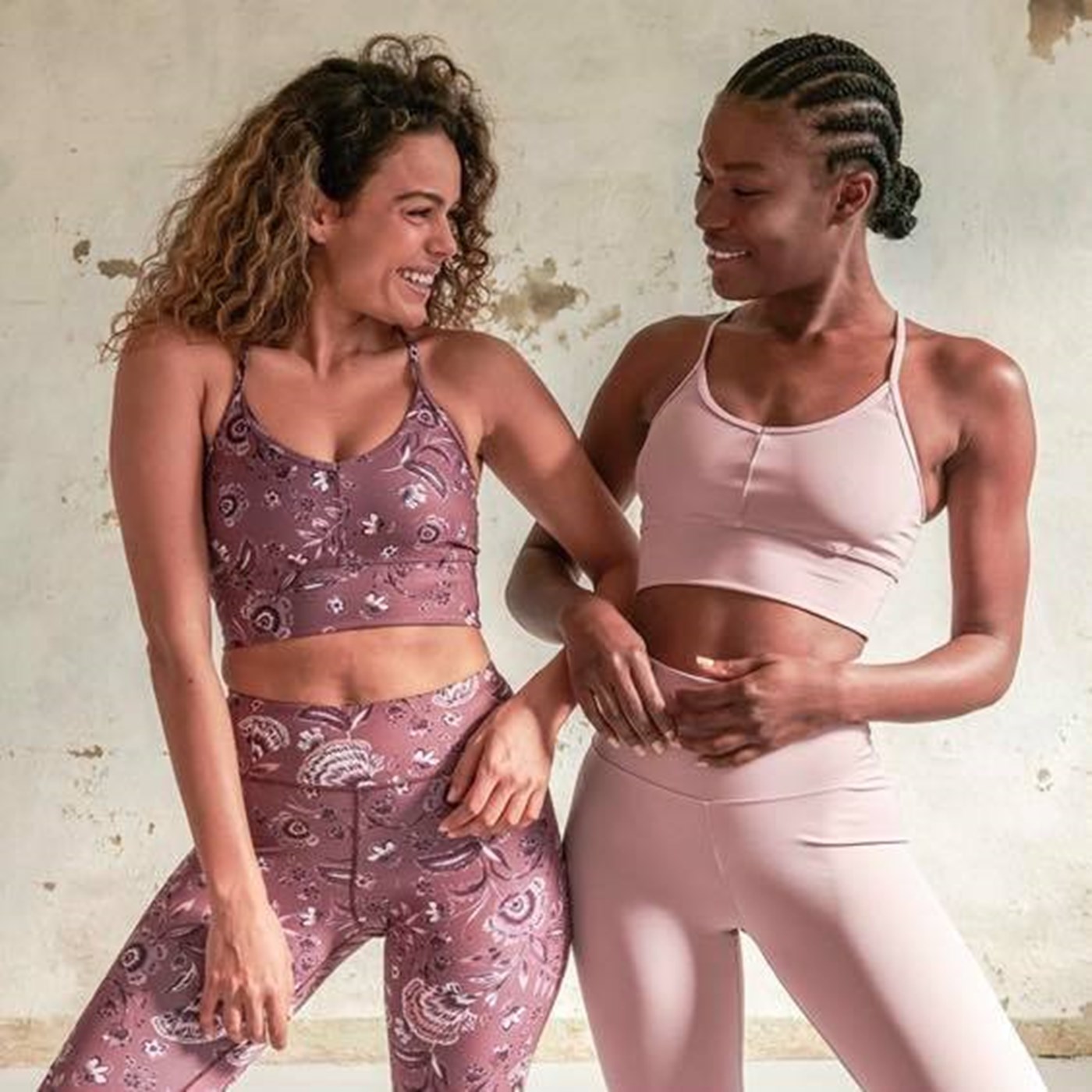 Two women in matching pink activewear looks link arms. 