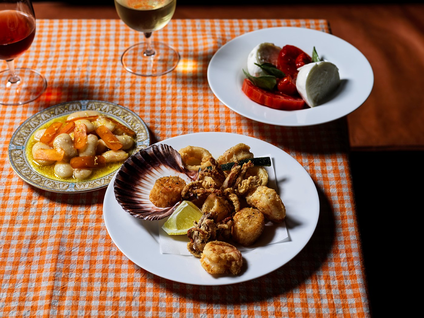 Plates of seafood, beans and fresh tomatoes and mozzarella against an orange and white gingham tablecloth 