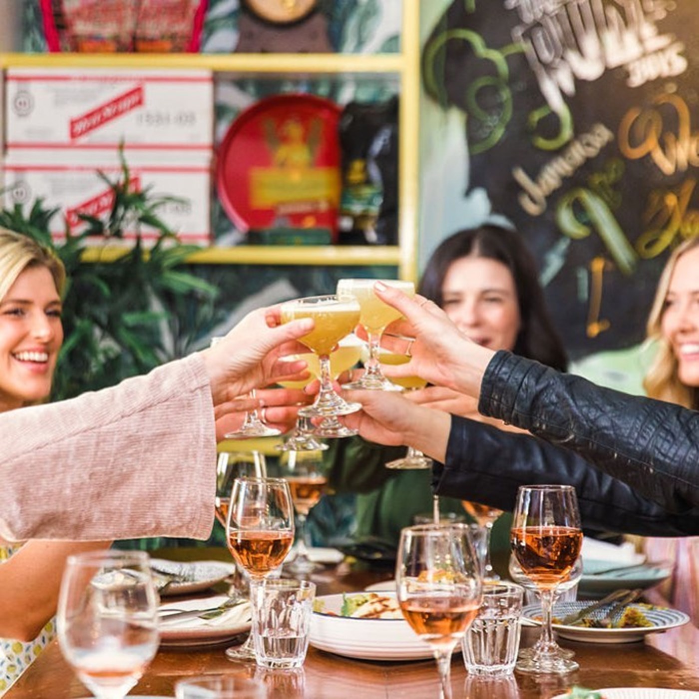 Multiple women leaning in over a table of wine to cheers with a cocktail 