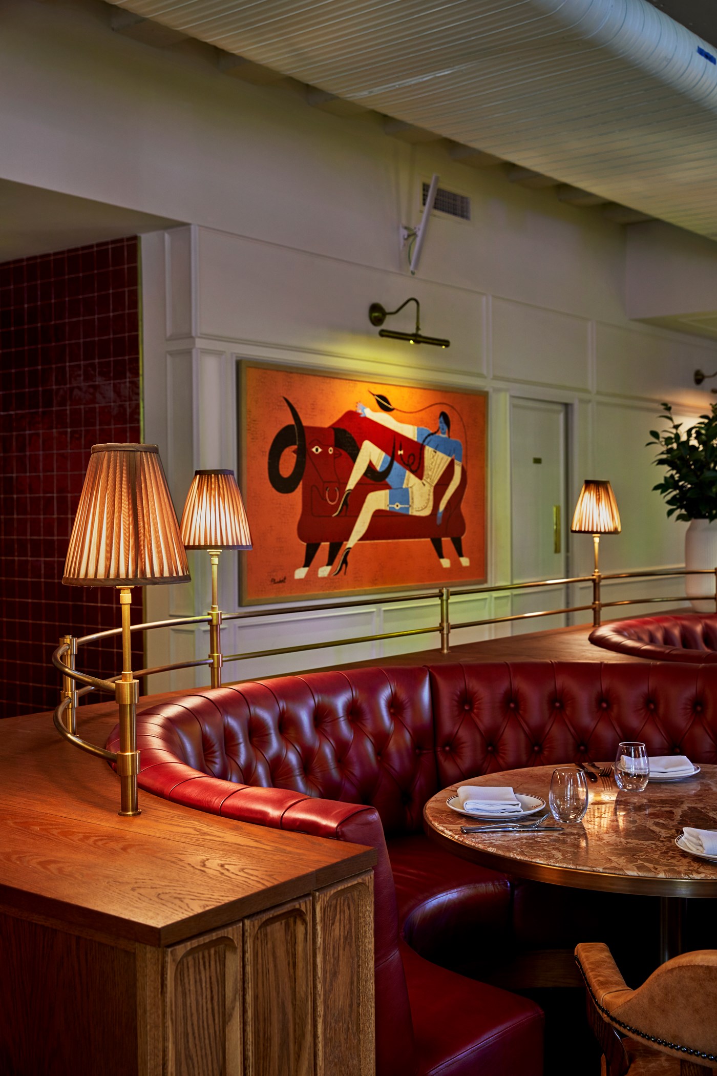A red leather corner booth, marble table and French lamps in front of a vibrant piece of modern artwork 
