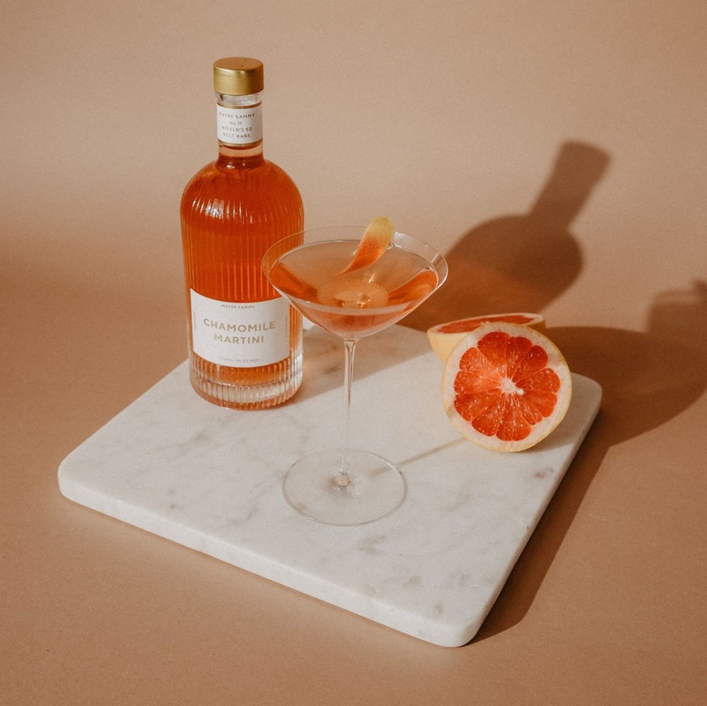 A bottled chamomile martini on a marble tray alongside a decanted martini and a cut open blood orange