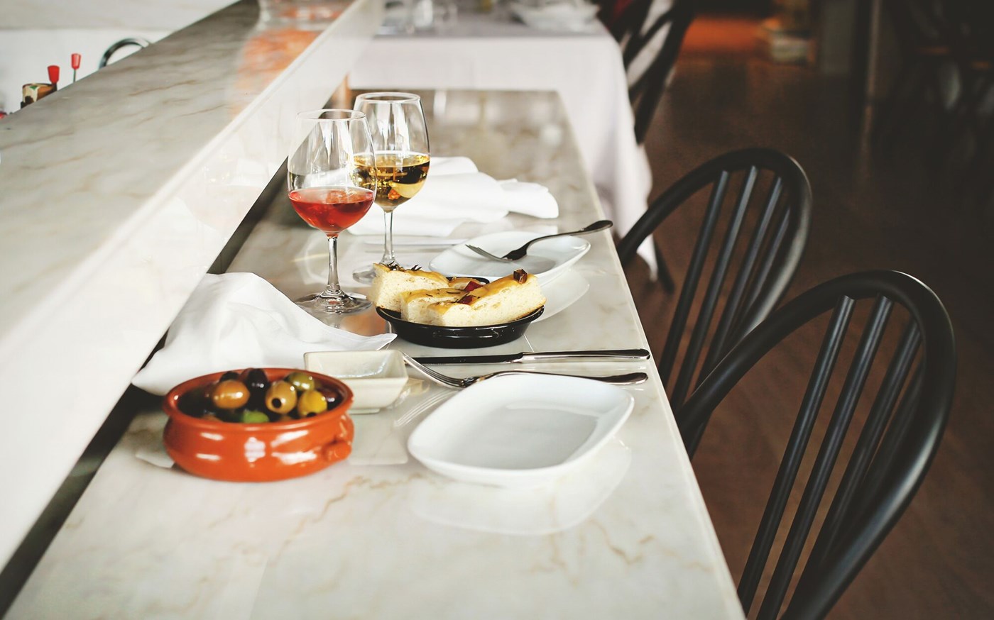 Two bar seats with a marble table filled on which sits olives, two glasses of wine and focaccia 