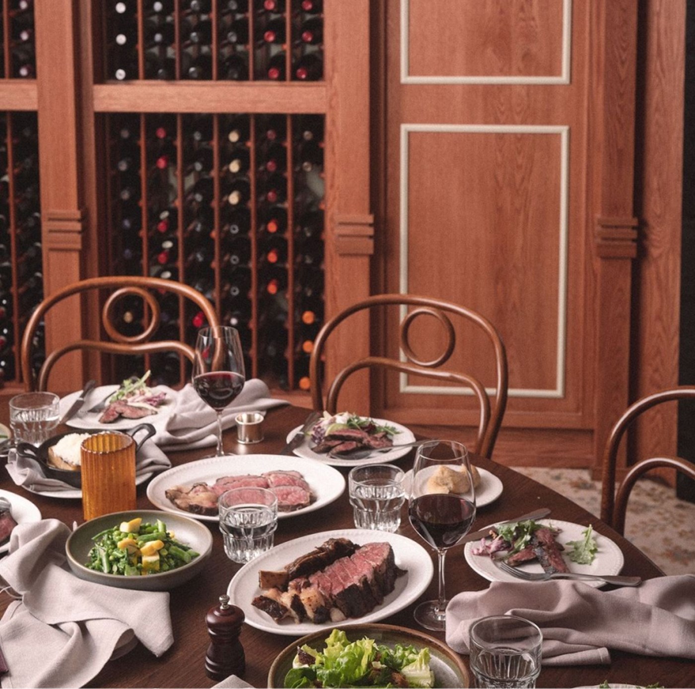 The edge of a circular table with plates of steak and salad against a backdrop of stacked wines 