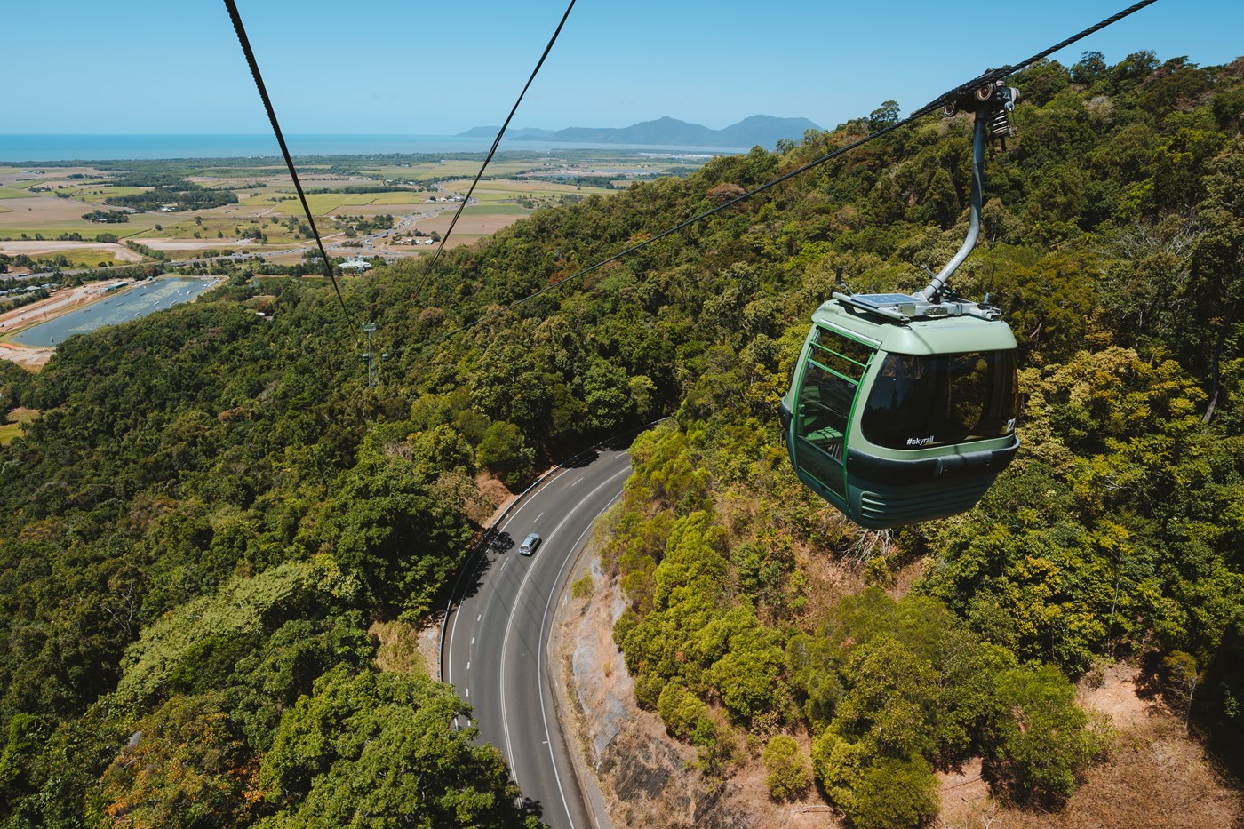 A cable car over a dense rainforest and a winding road beneath it