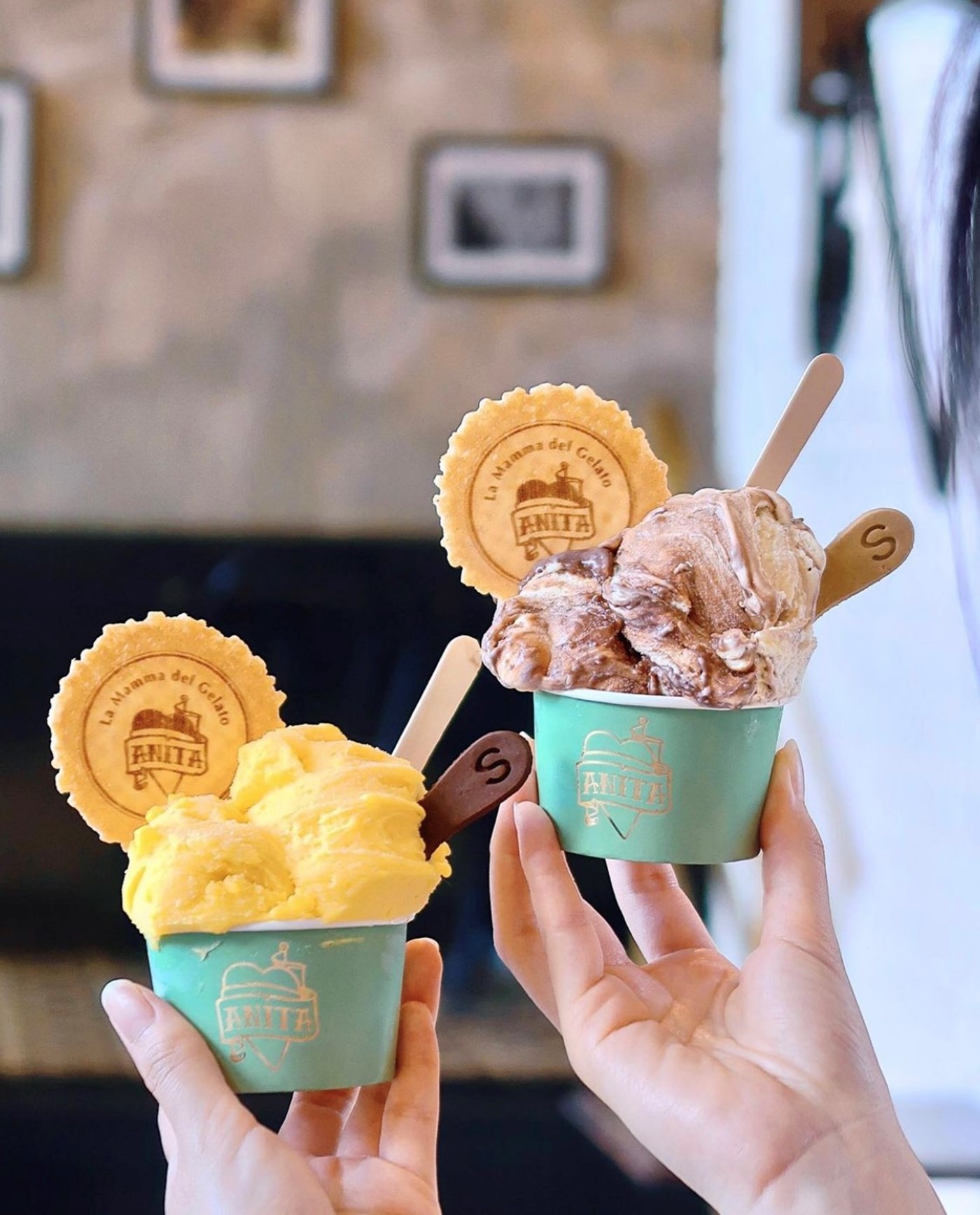 Two hands display two flavours of gelato in cups, one scoop of mango and one scoop of cookies and cream.