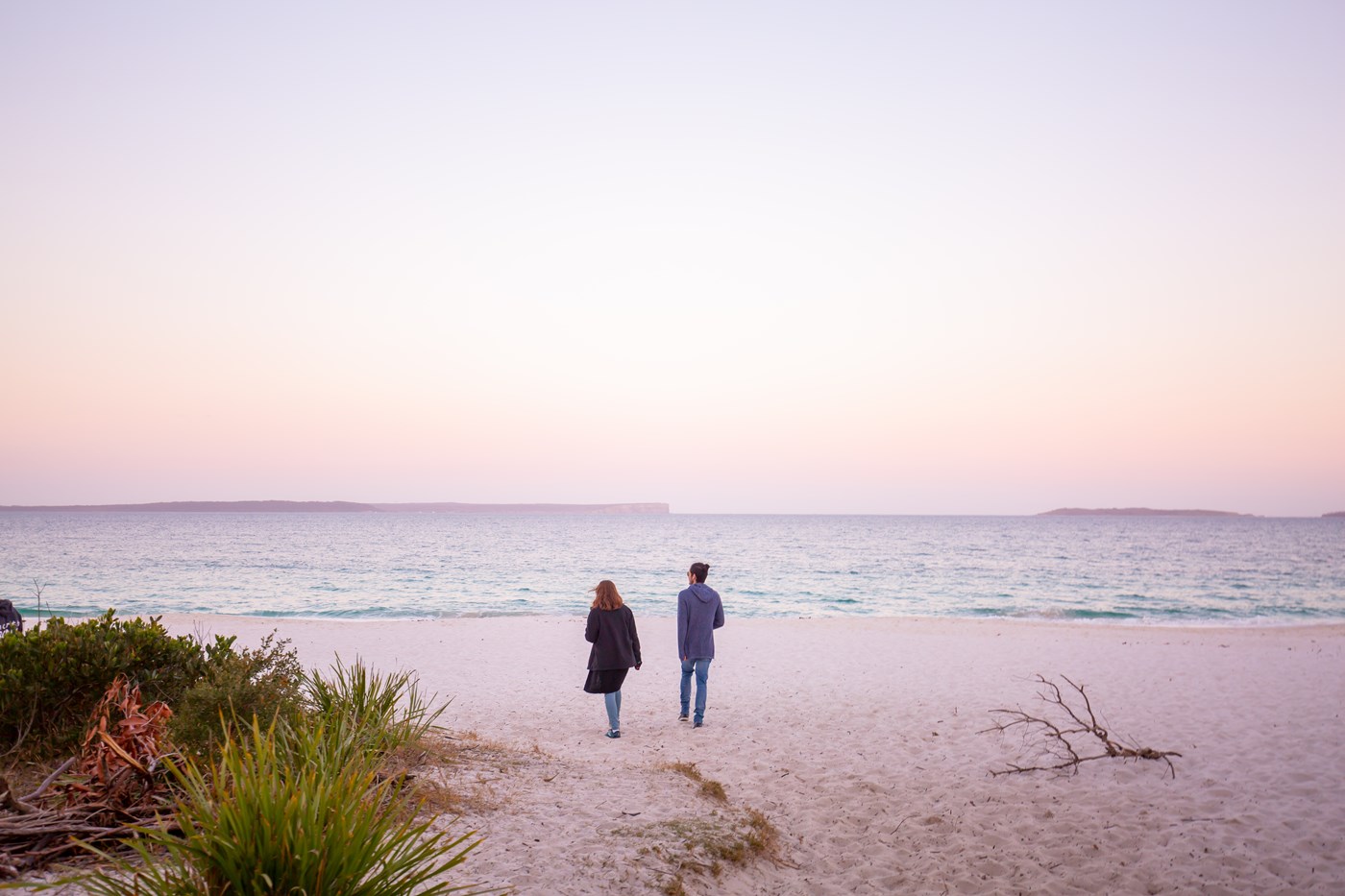 A man and woman walk on the beach towards the ocean during sunset at Jervis Bay NSW