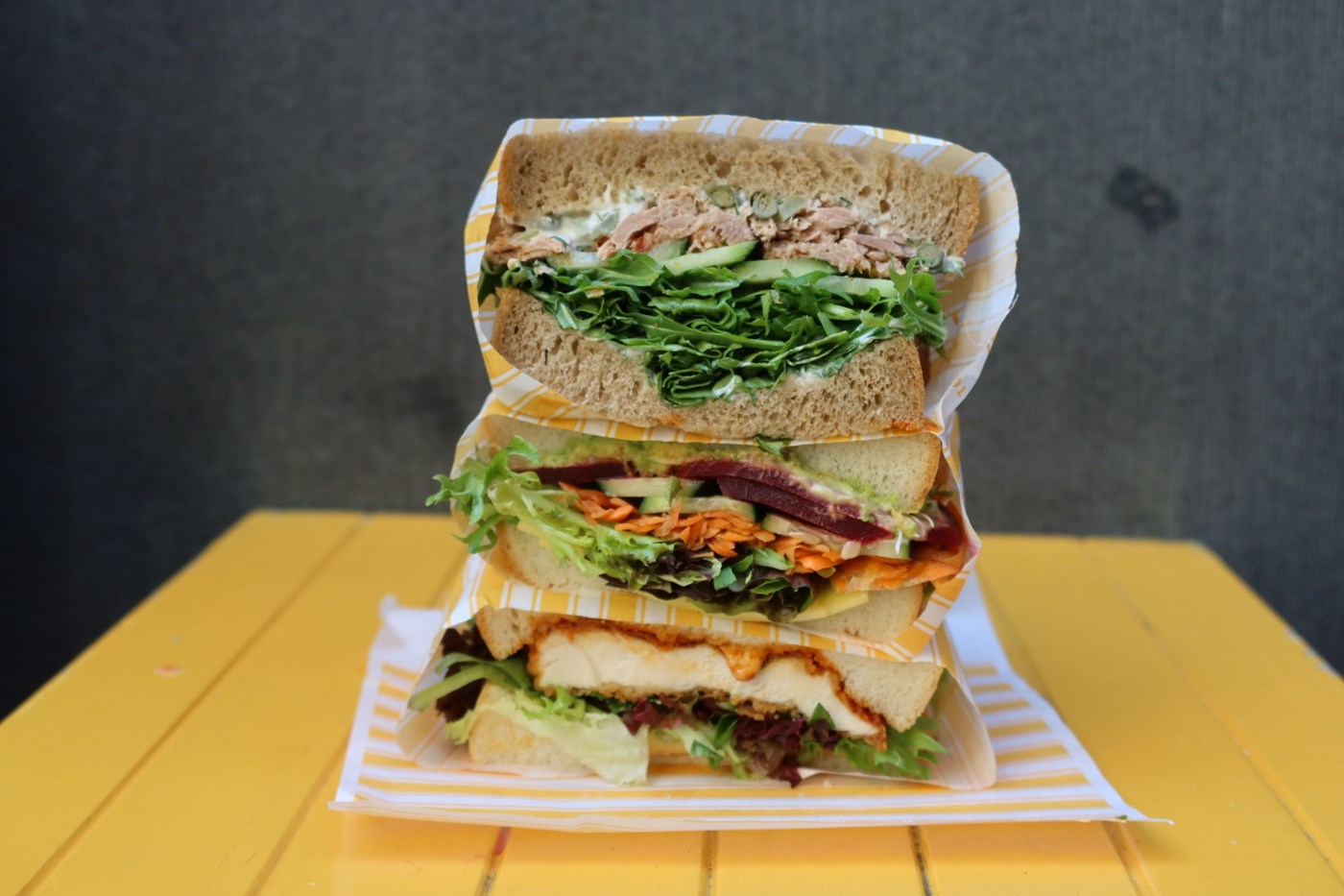 A stack of three sandwiches on a yellow wooden table by Junes Sandwich Shoppe