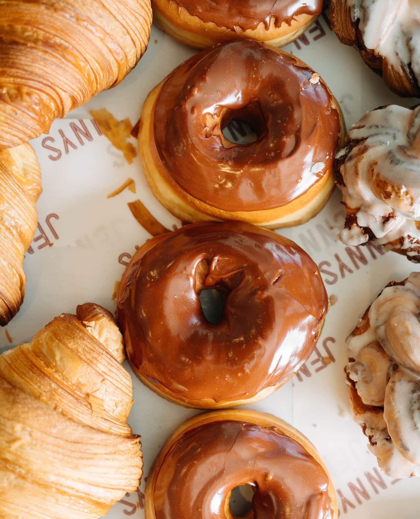 Chocolate-glazed donuts alongside croissants and iced cinnamon rolls on branded paper at Jenny's Bakery, Adelaide.