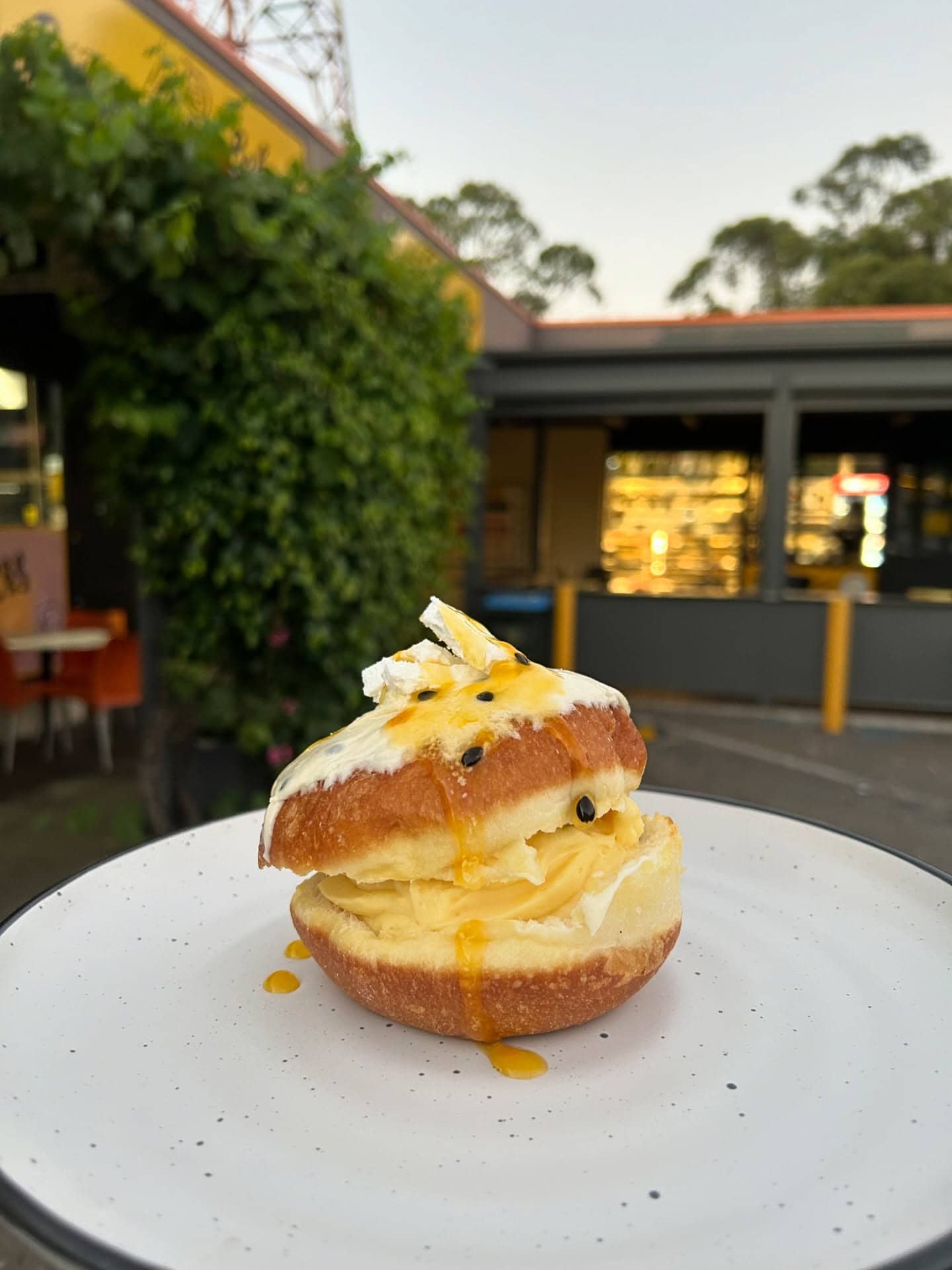 A cream-filled donut topped with passionfruit curd and whipped cream on a white plate outside Banana Boogie Bakery, Adelaide, at dusk.