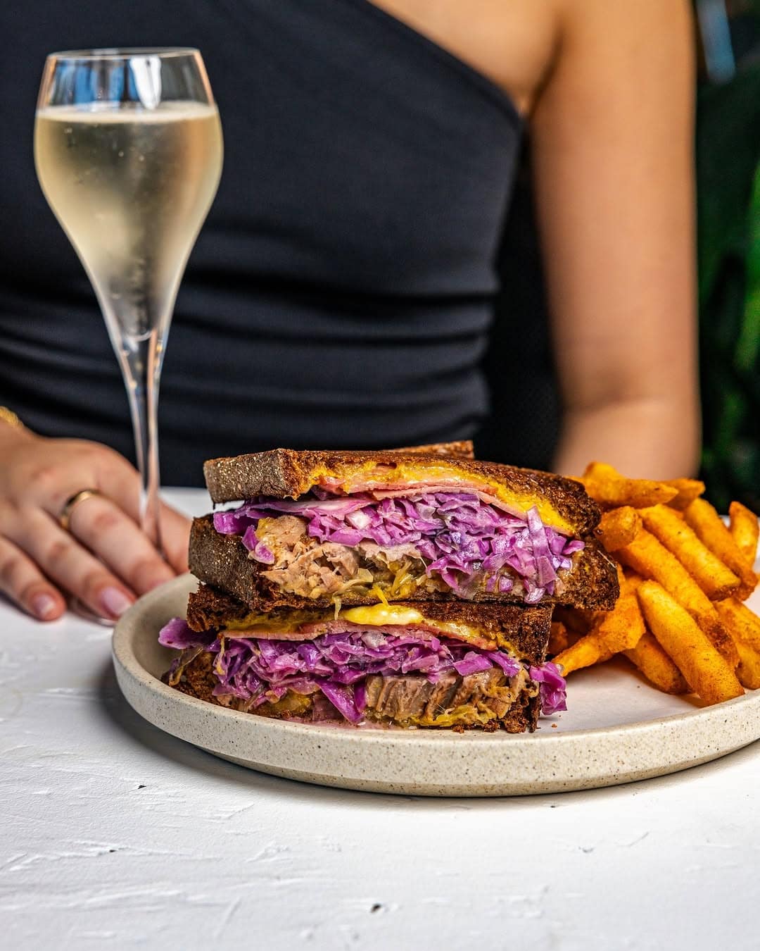 A woman holds a glass of champagne with a burger and fries on the table.