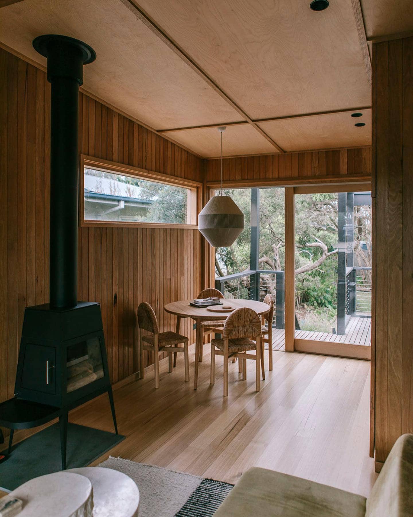 Light-filled dining area at YOKO Luxury Cabin, featuring timber interiors, a freestanding wood-burning fireplace and glass doors opening onto a private deck.