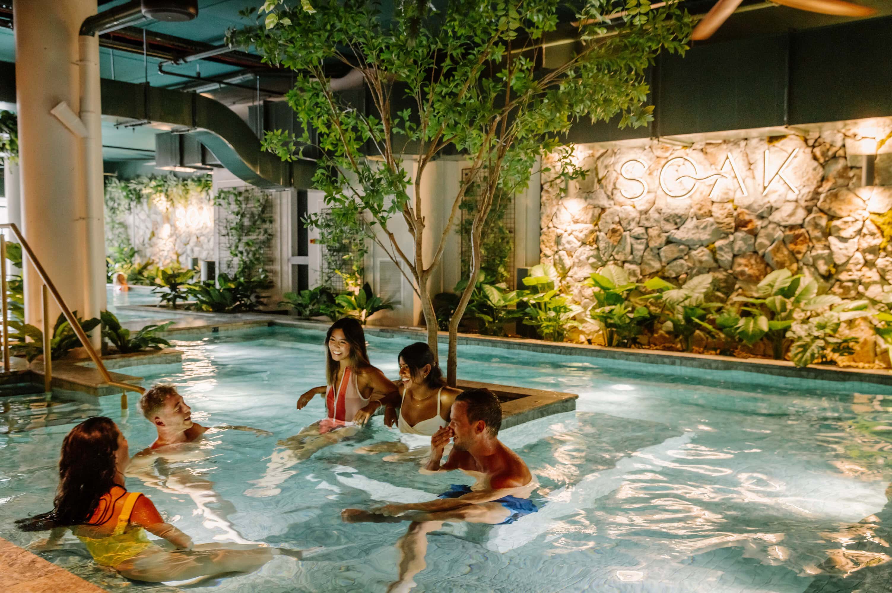 Friends laughing in magnesium pool with neon Soak sign and greenery at Soak Bathhouse Sydney