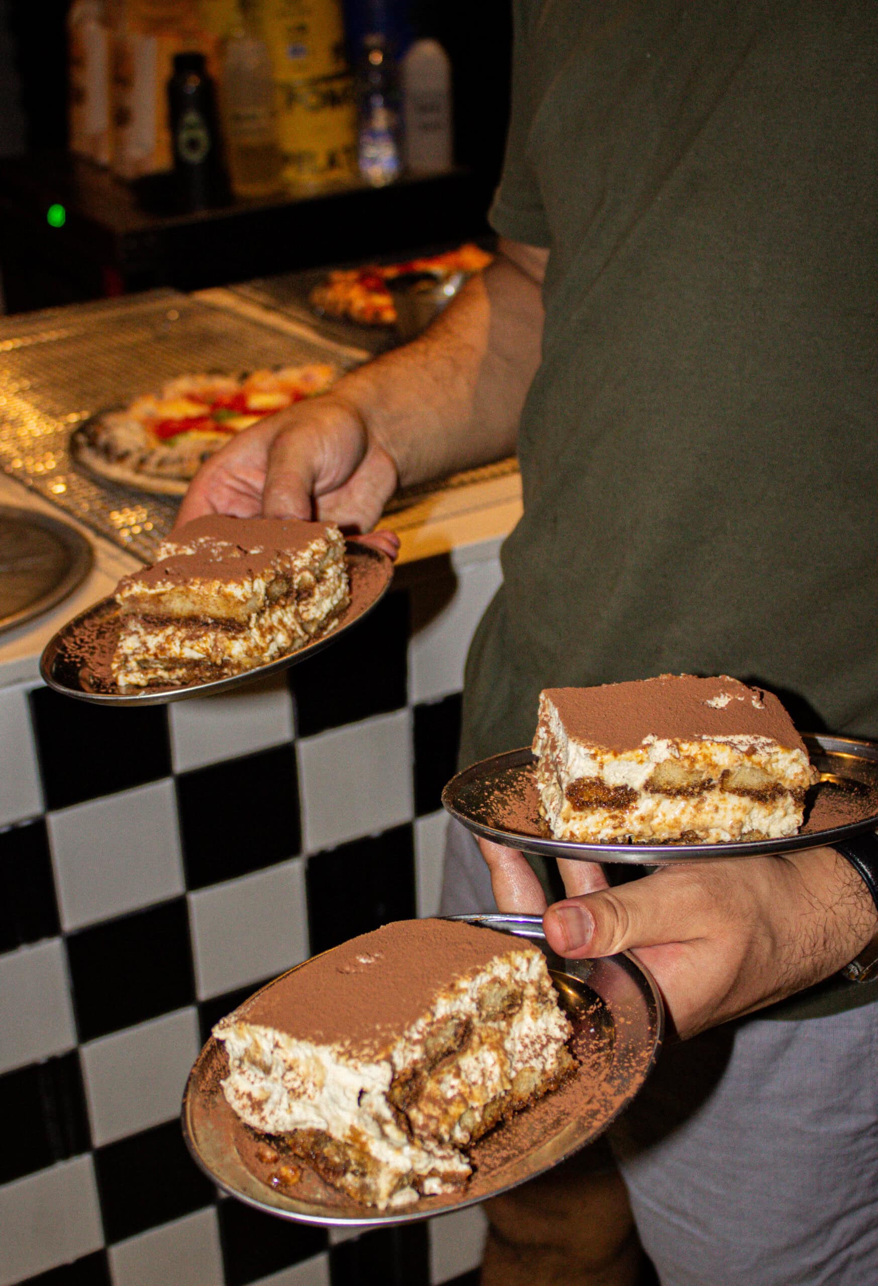 Plates of tiramisu carried past a black and white chequered tile counter at Pizzeria Officina, Braddon Canberra