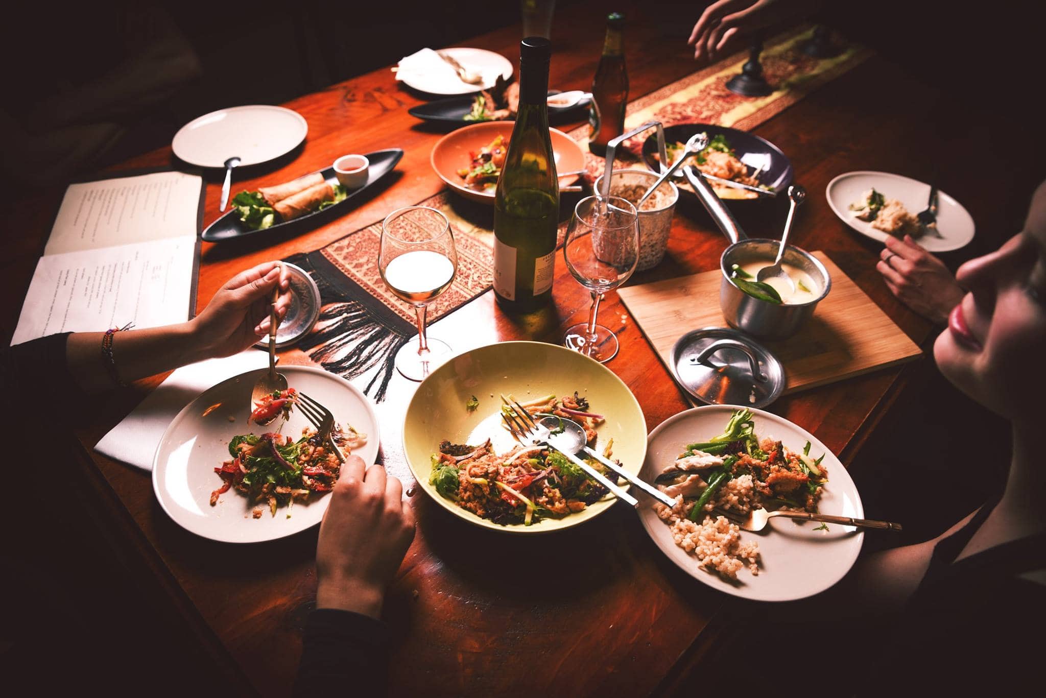 Friends sharing a Thai banquet at Sabai Cafe, Adelaide, with an array of dishes including salads, stir-fries, rice, and soup spread across the restaurant's signature dark timber table, with white wine and warm ambient lighting.