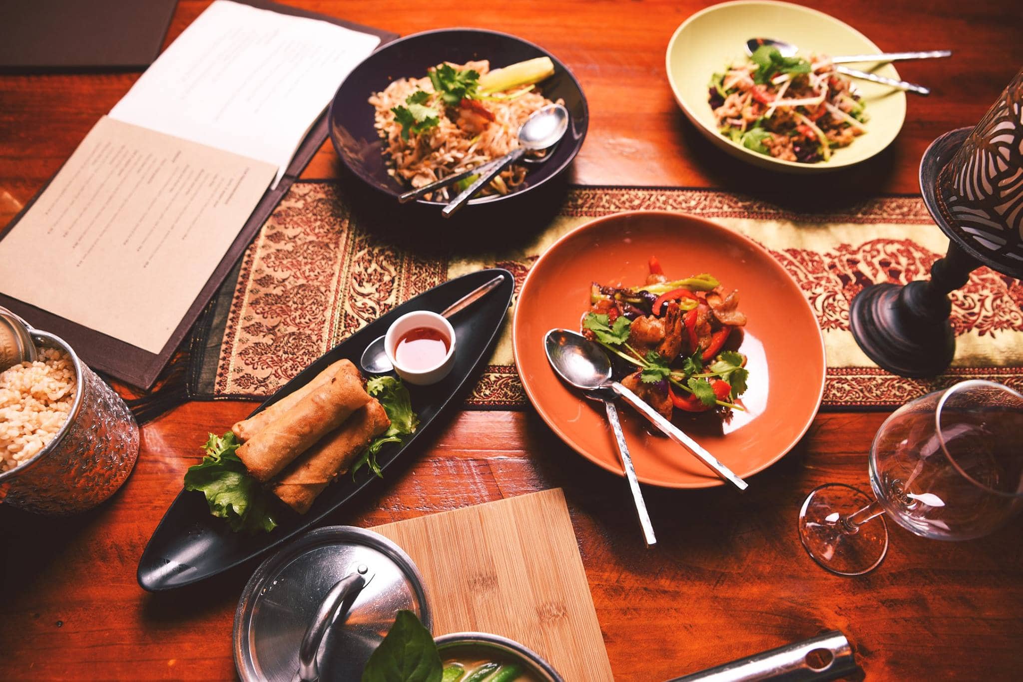 Overhead view of a Thai feast at Sabai Cafe set on the restaurant's warmly lit timber table with a Sabai menu in Adelaide.