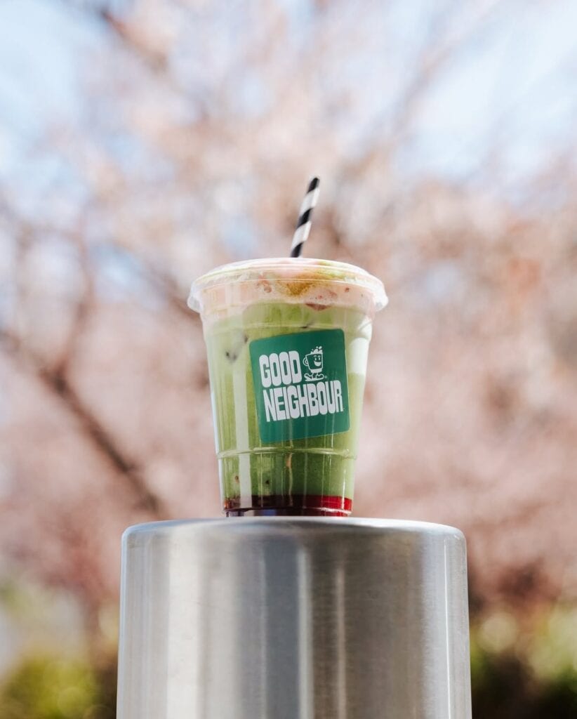 Good Neighbour iced layered matcha drink in branded cup with striped straw against cherry blossom background Kingston Canberra