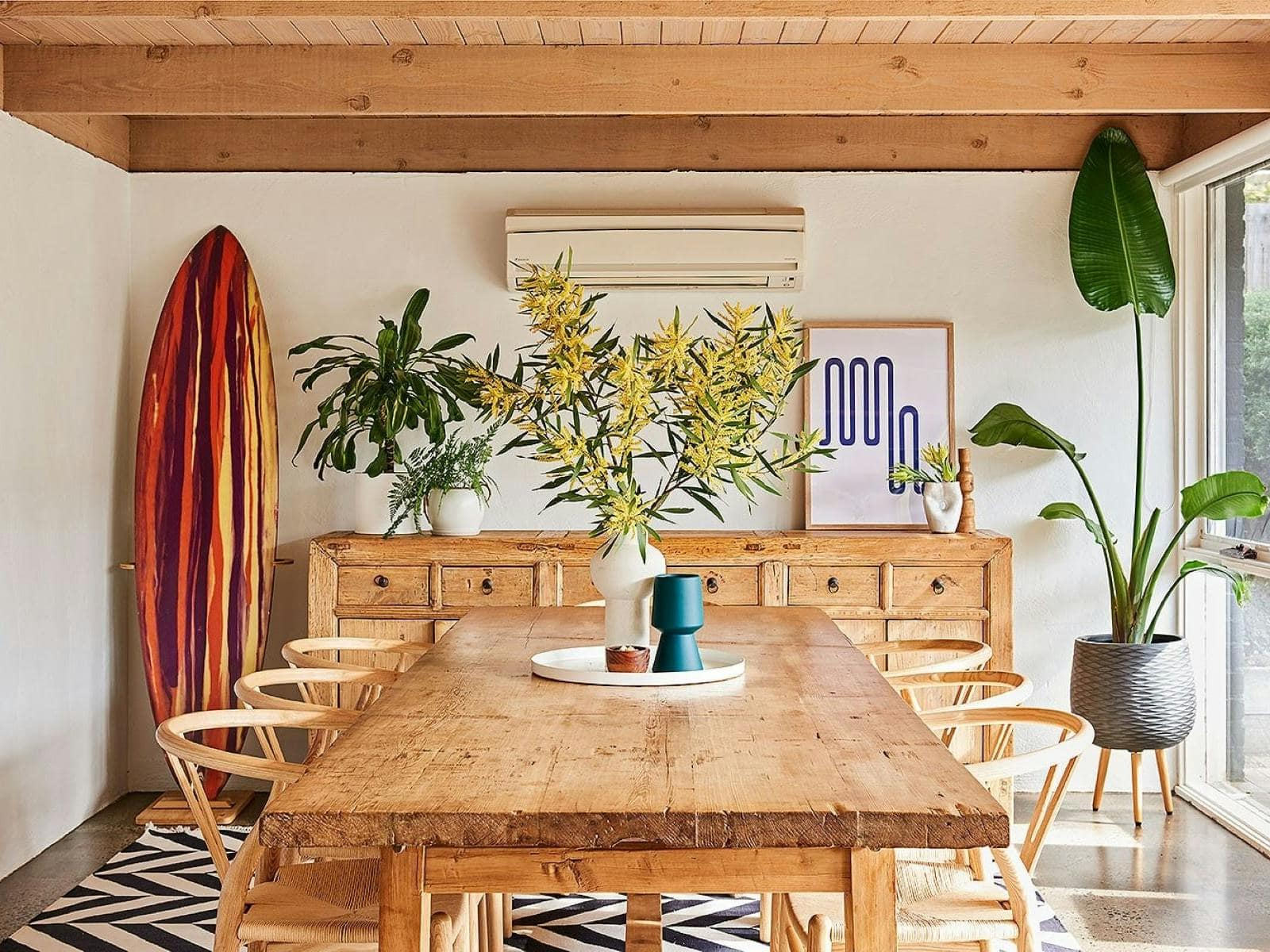 Dining room at 100 Steps to the Beach in Anglesea, with a long timber table, surfboard, indoor plants and natural light.